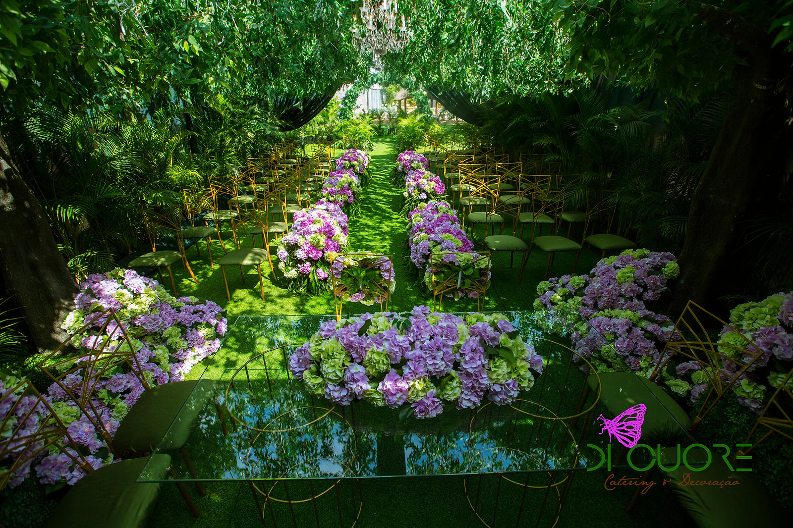 Glass table heaped with lilac and lime-green hydrangeas, with the aisle of gold chairs and hanging foliage beyond