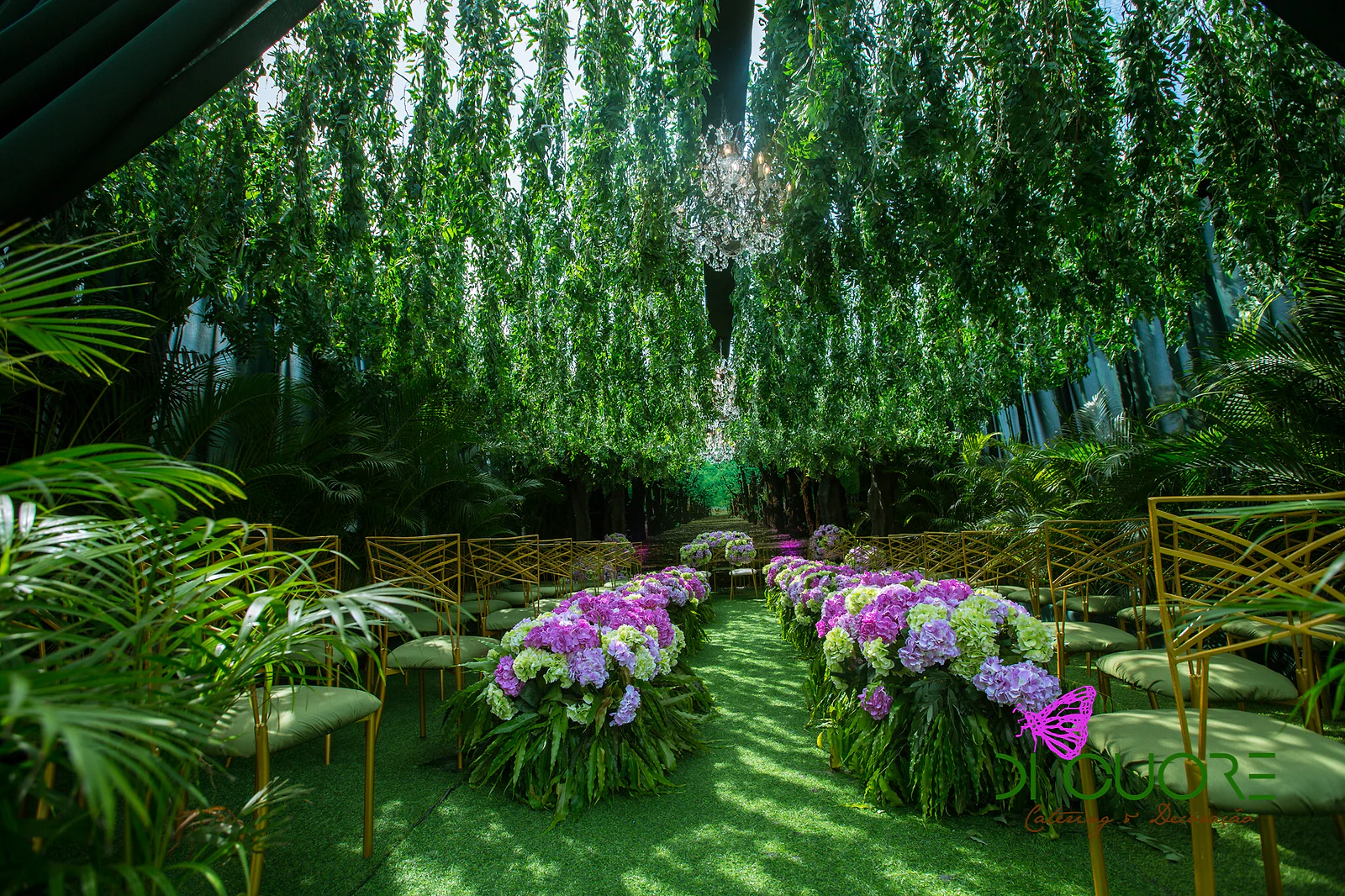Grass aisle between rows of hydrangeas and gold chairs, beneath hanging sprays of foliage and a crystal chandelier