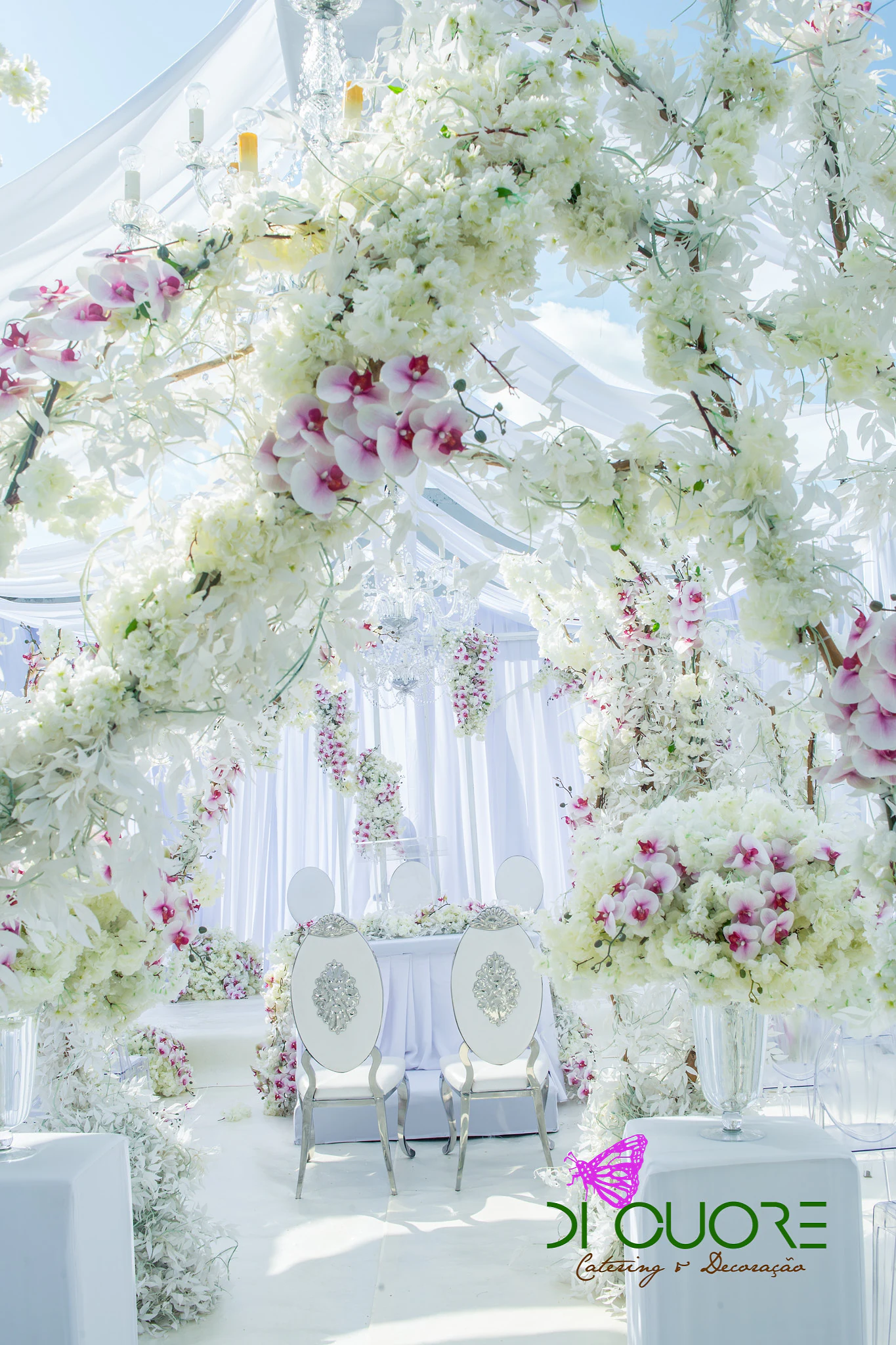 Arches of white flowering branches with pale pink orchids above the couple’s chairs, between white drapes