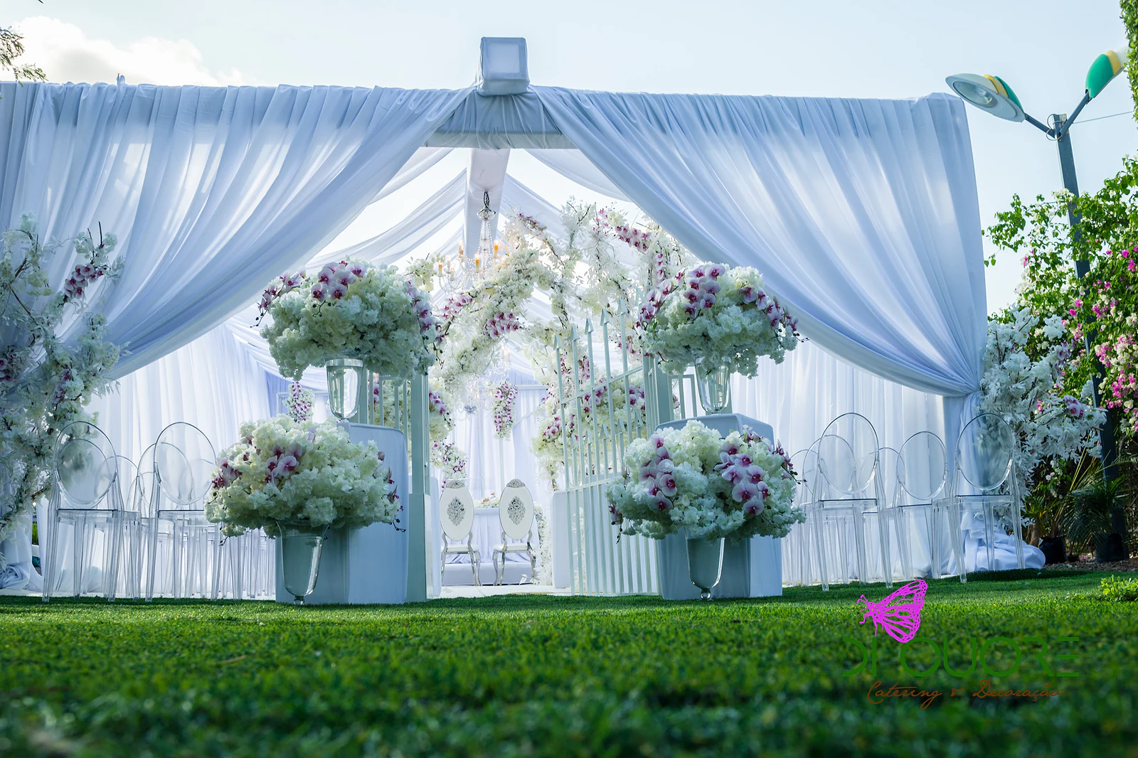 White-draped pavilion open onto the lawn, with tall vases of white flowers and orchids and clear chairs inside