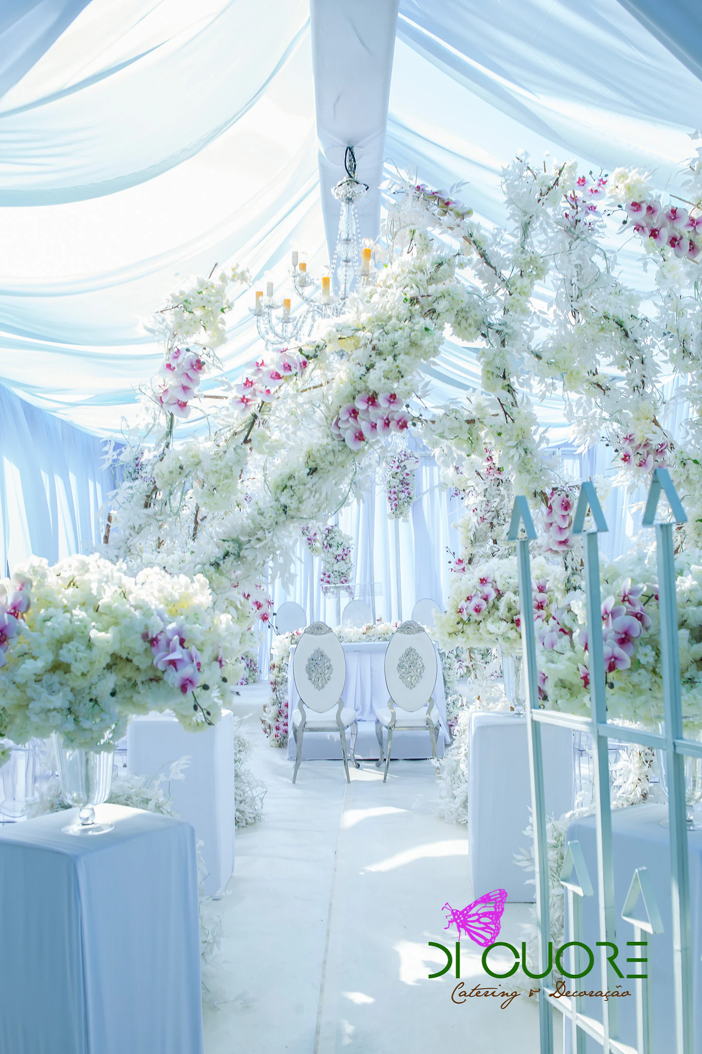 White aisle with arches of flowering branches and orchids leading to the couple’s chairs, beneath pale blue drapes