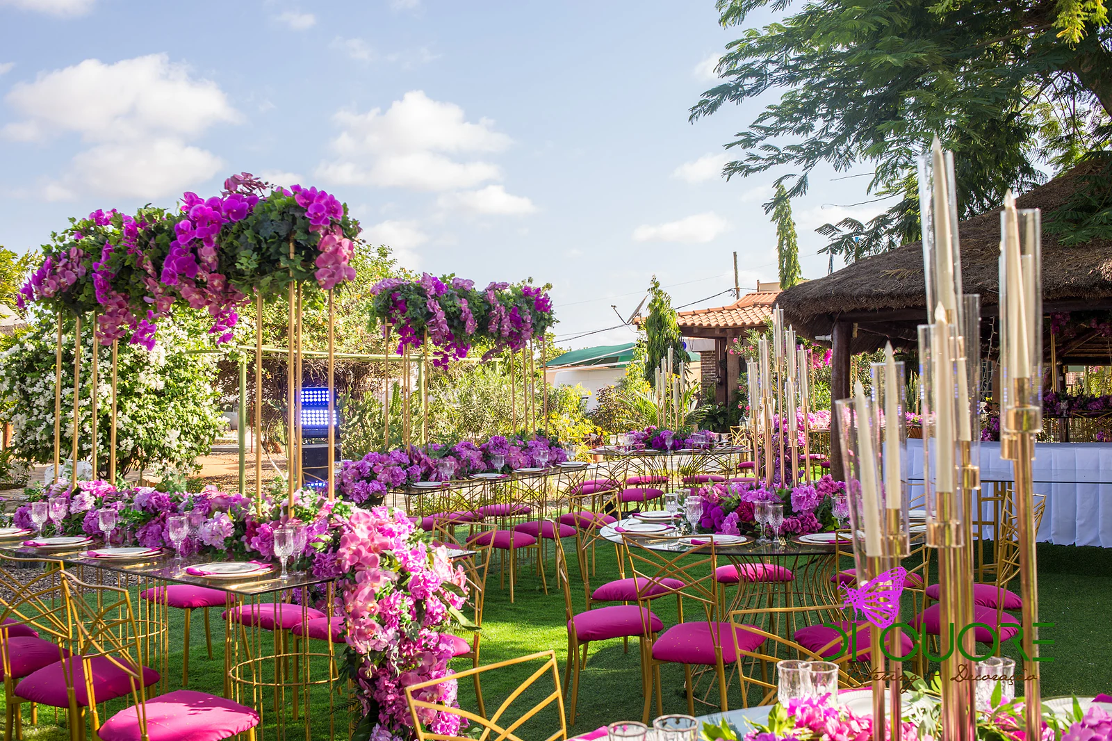 Glass tables with orchid centrepieces, tall gold candlesticks and chairs with fuchsia cushions, in an open-air garden