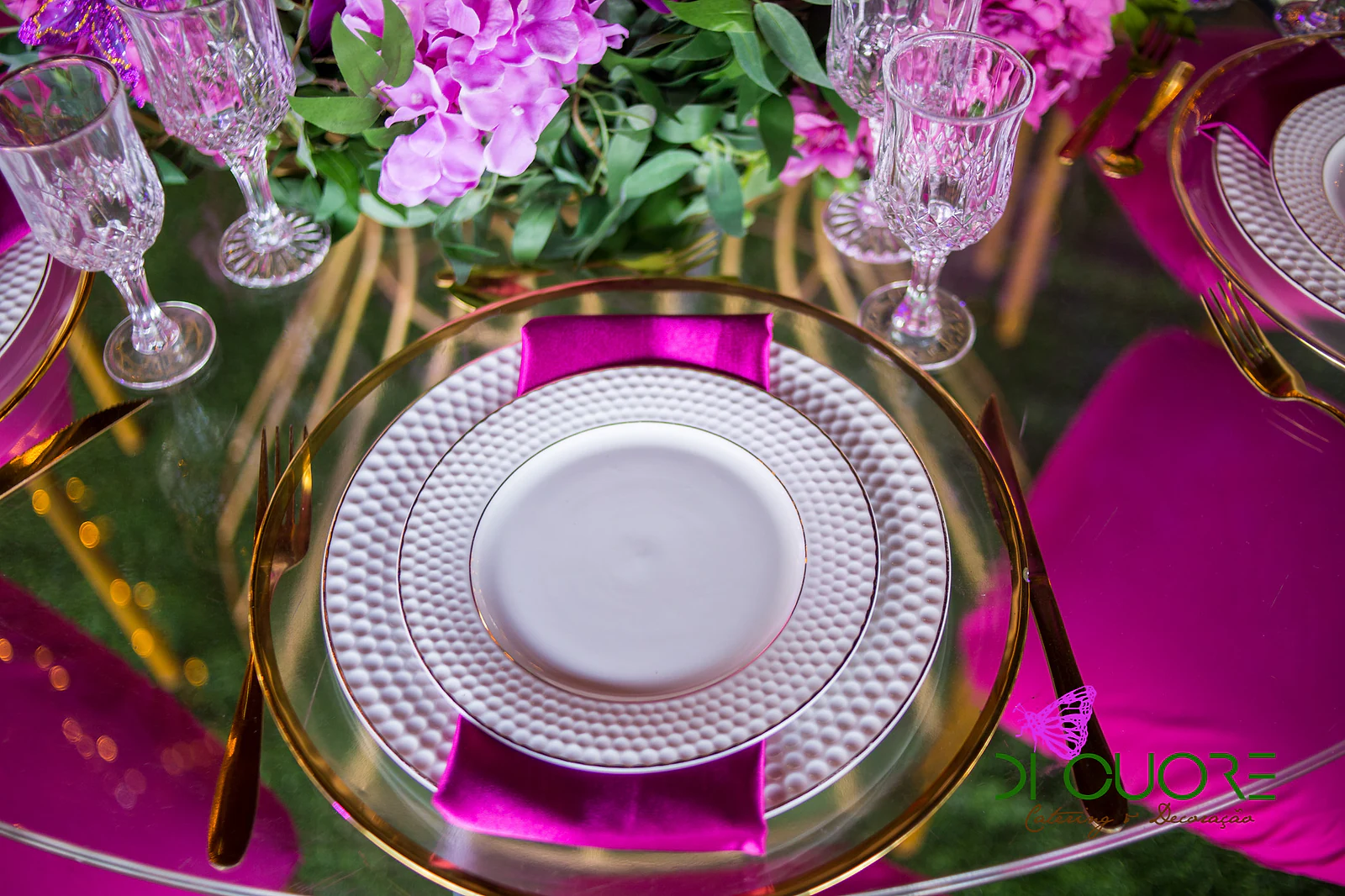 Place setting with a textured white plate on a gold charger, a fuchsia napkin and crystal glasses, among pink orchids