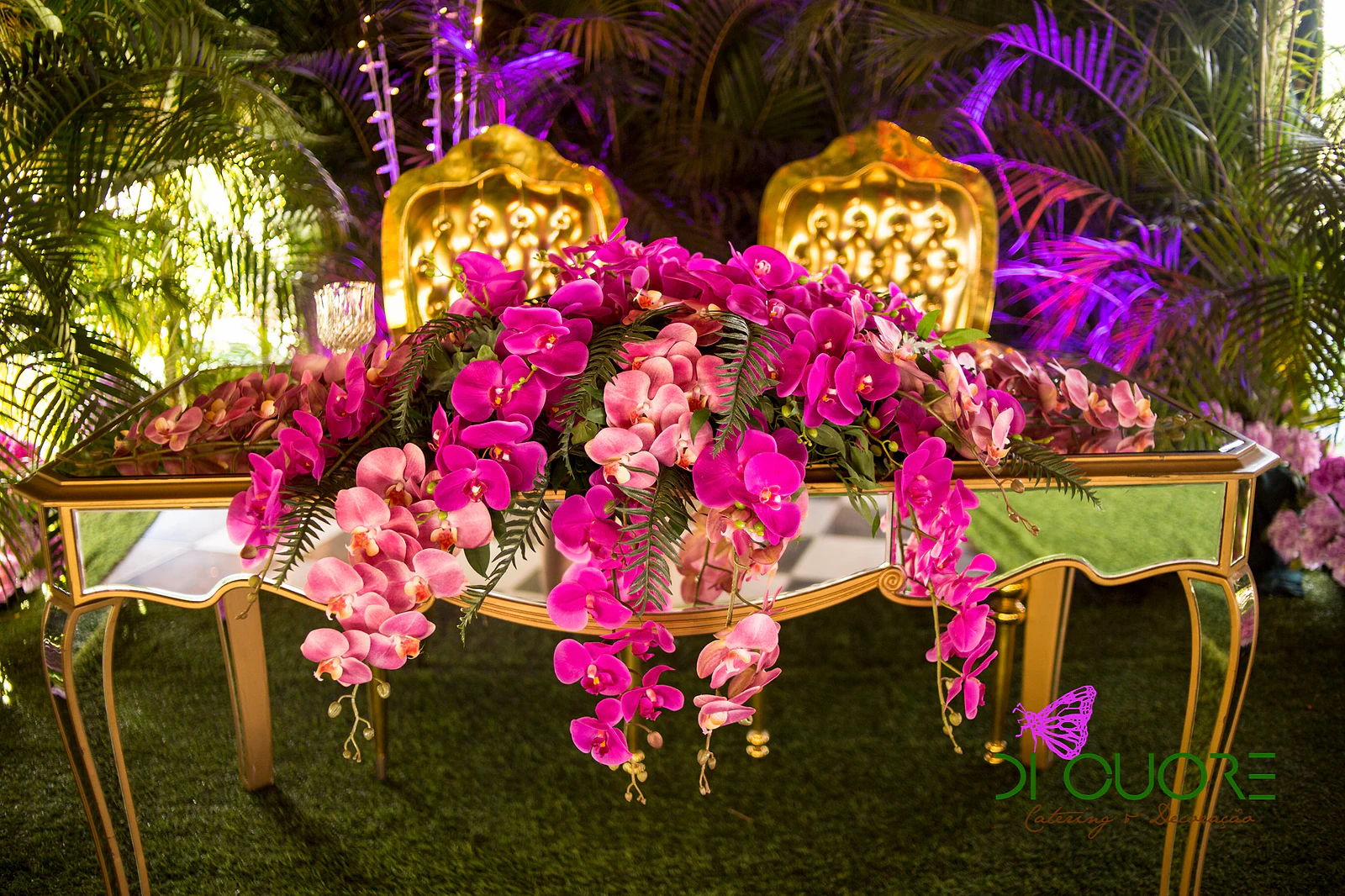 Mirrored and gold table with a large arrangement of pink and fuchsia orchids, in front of two gold chairs, with purple light beyond