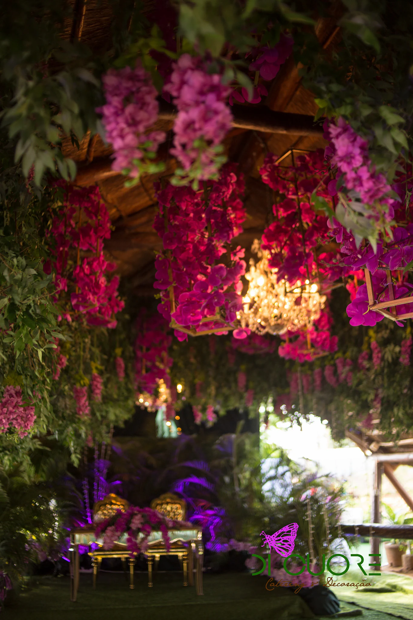 Thatched ceiling covered in hanging orchids and foliage, with a lit crystal chandelier and the couple’s table beyond, in purple light