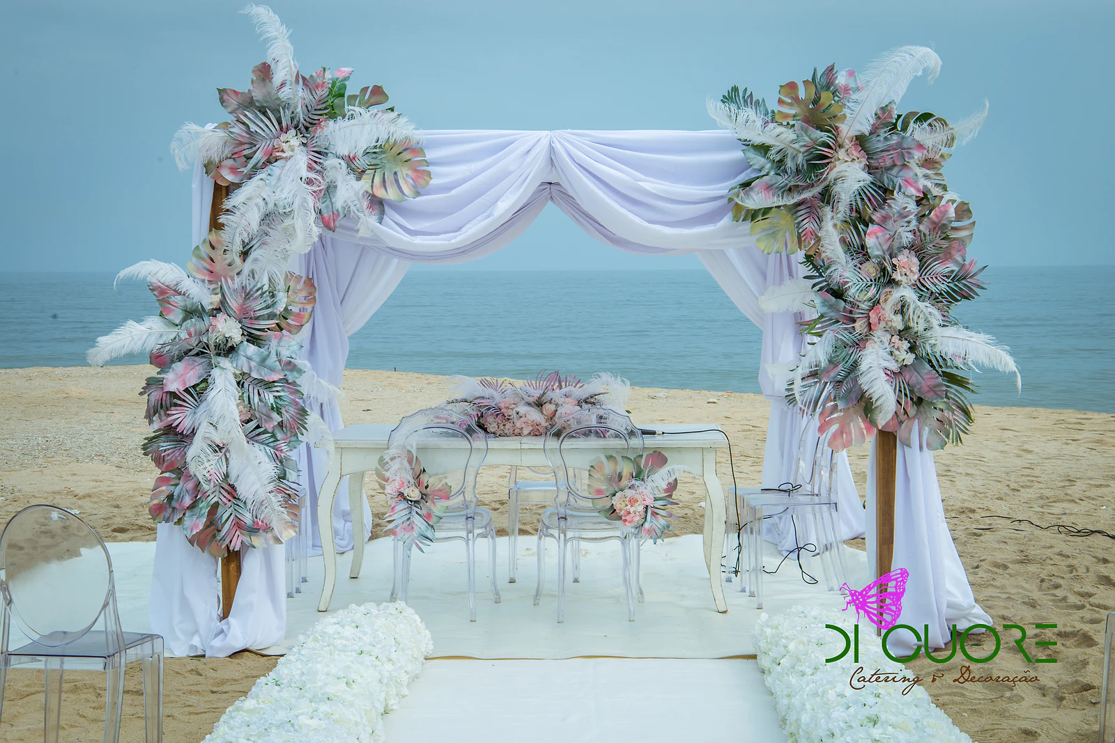 Dossel de tecido branco drapeado sobre a mesa dos noivos, entre duas colunas de folhas rosa e verde e penas brancas, com o mar ao fundo