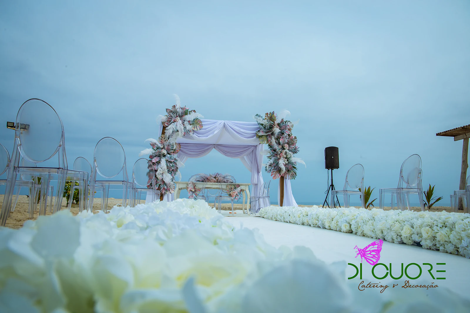 Cadeiras transparentes na areia ao longo da passadeira e de uma fiada de flores brancas, com hortênsias em primeiro plano e o dossel ao longe
