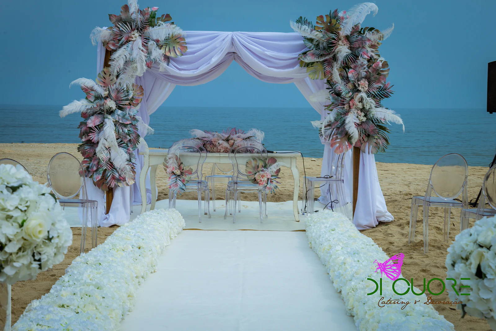 White carpet between rows of white hydrangeas leading to the draped fabric canopy, with columns of leaves and feathers and the sea beyond