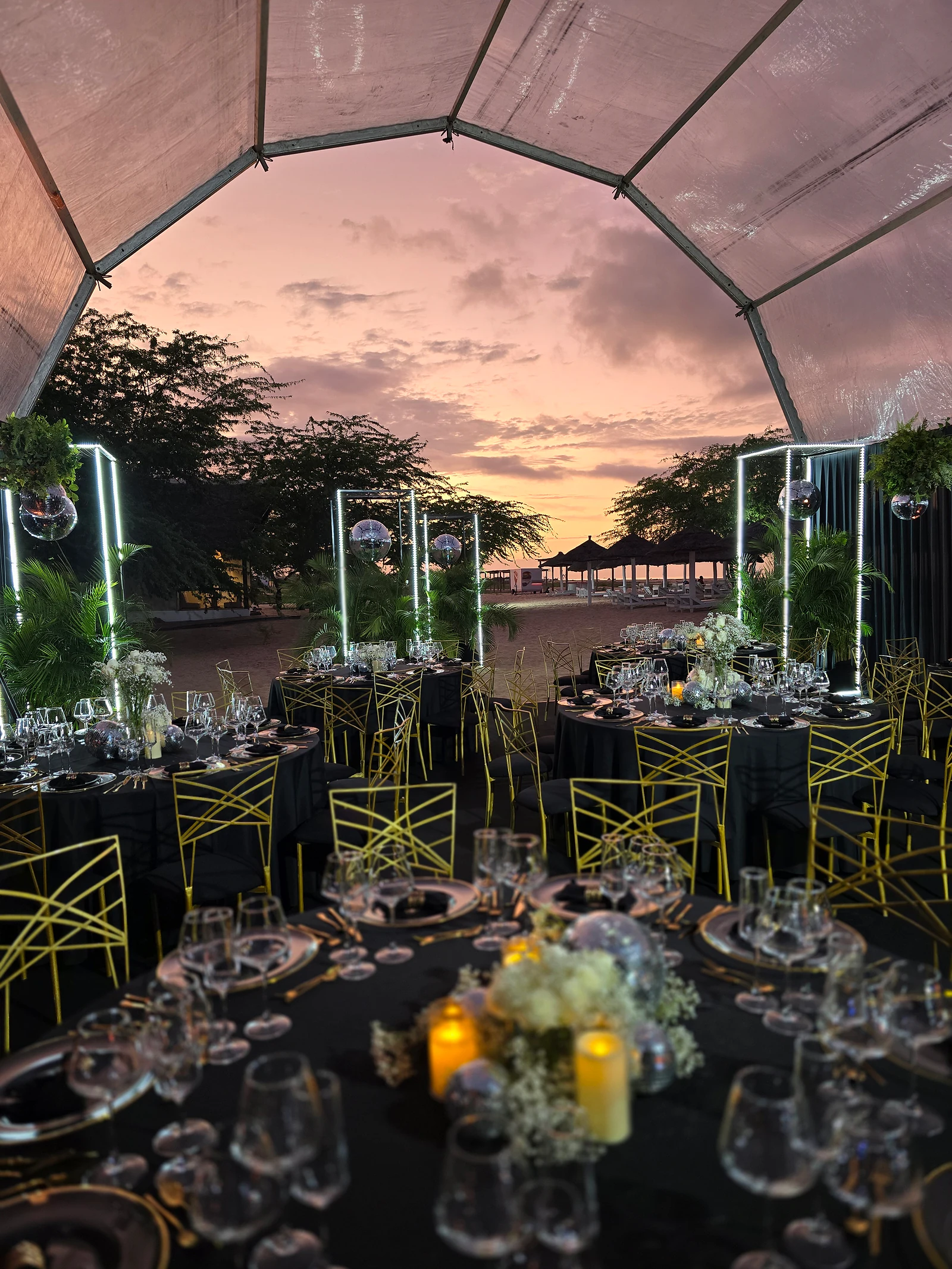 Mesa redonda em primeiro plano com velas acesas e um centro de flores brancas e, ao fundo, a tenda aberta ao céu cor-de-rosa do entardecer