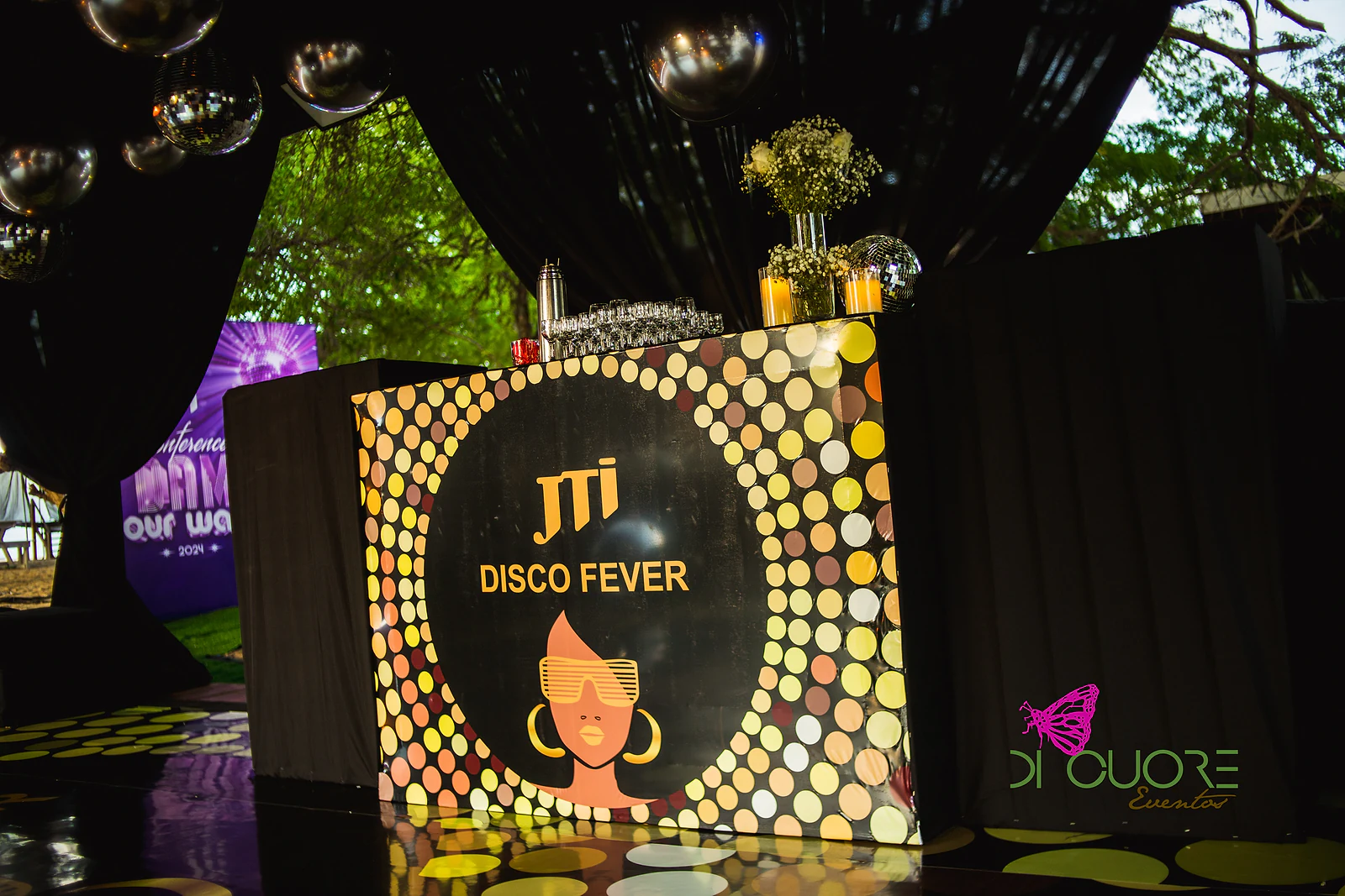 Balcão preto com o logótipo JTI Disco Fever num círculo de pontos dourados, com um ramo de gipsófila ao cimo