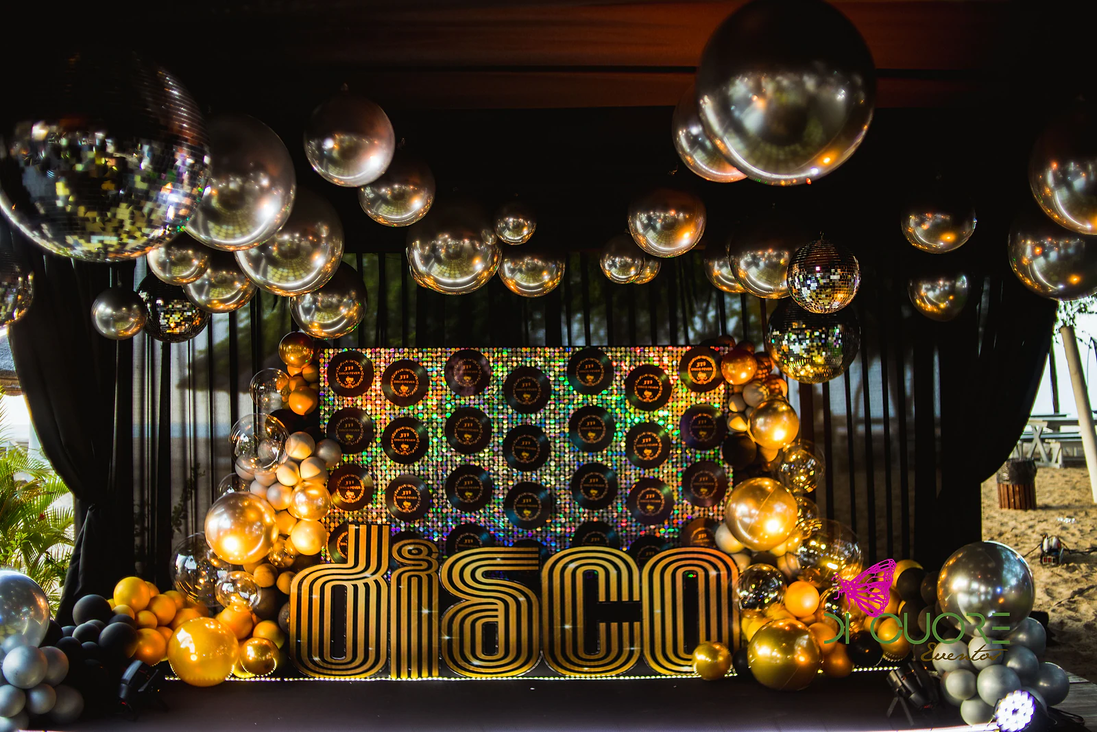 Palco com a palavra DISCO em letras douradas diante de um painel holográfico de vinis, rodeado de balões dourados e prateados e de esferas espelhadas no tecto