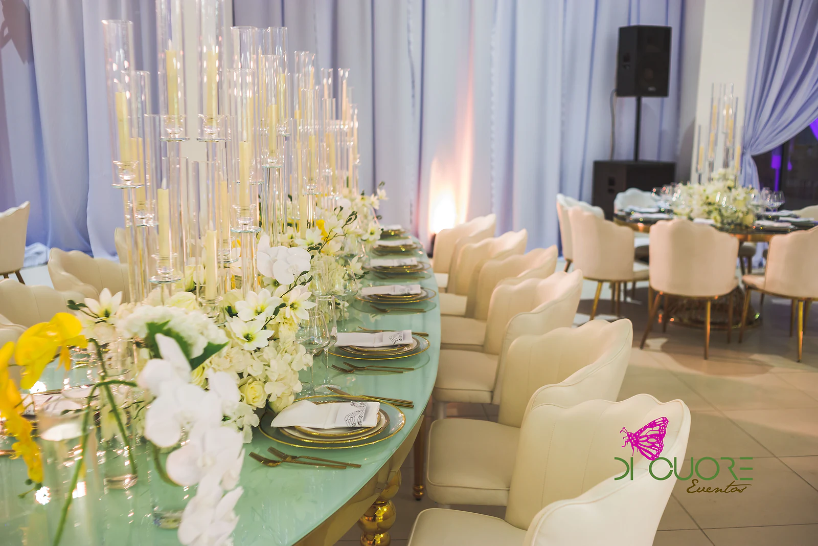 Mesa corrida de tampo verde-água com candelabros de velas em cilindros de vidro, orquídeas brancas e cadeiras creme