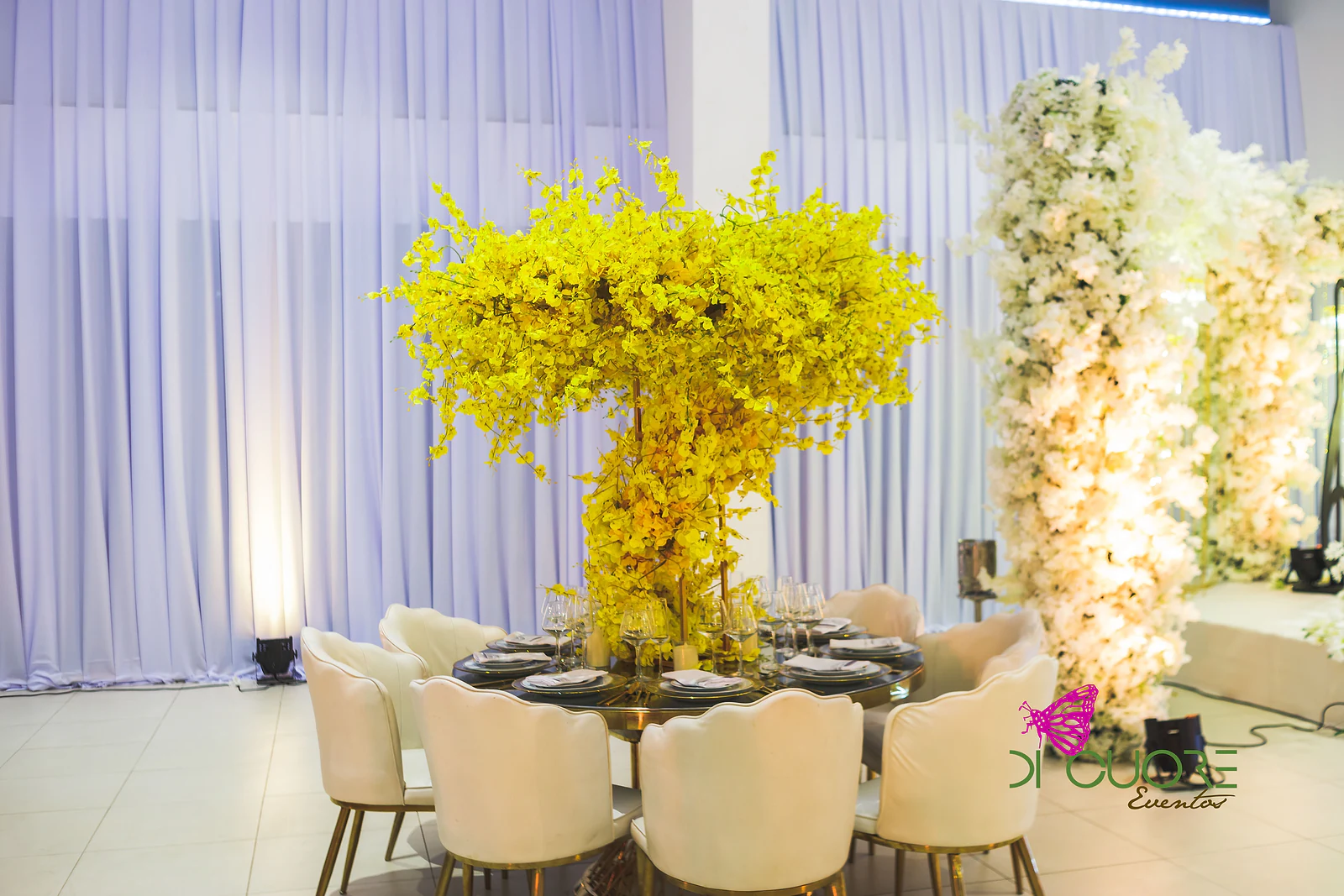 Mesa redonda com uma árvore de flores amarelas ao centro, cadeiras creme em forma de concha e uma coluna de flores brancas ao lado