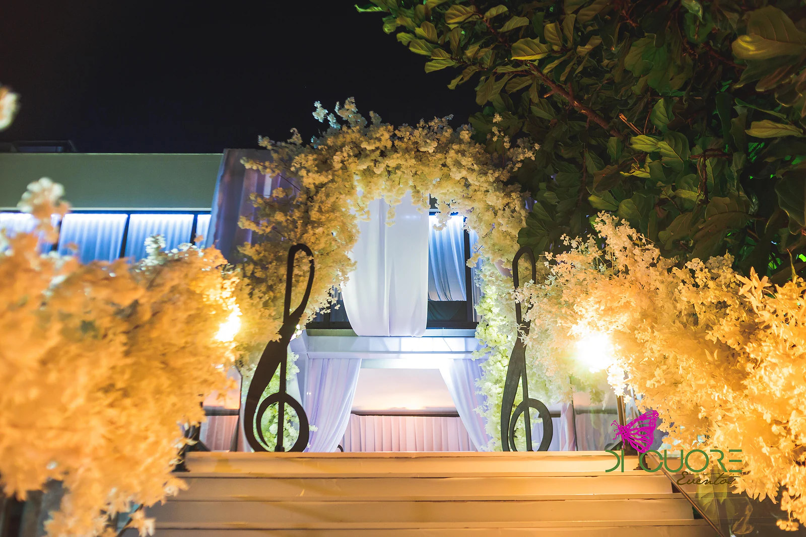 Escadas ao ar livre, de noite, com um arco de flores brancas e duas claves de sol iluminados por focos quentes