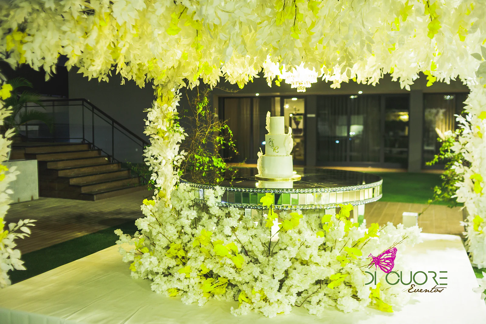 Bolo branco de vários andares sobre uma mesa espelhada, sob um dossel de flores brancas, junto a um jardim iluminado