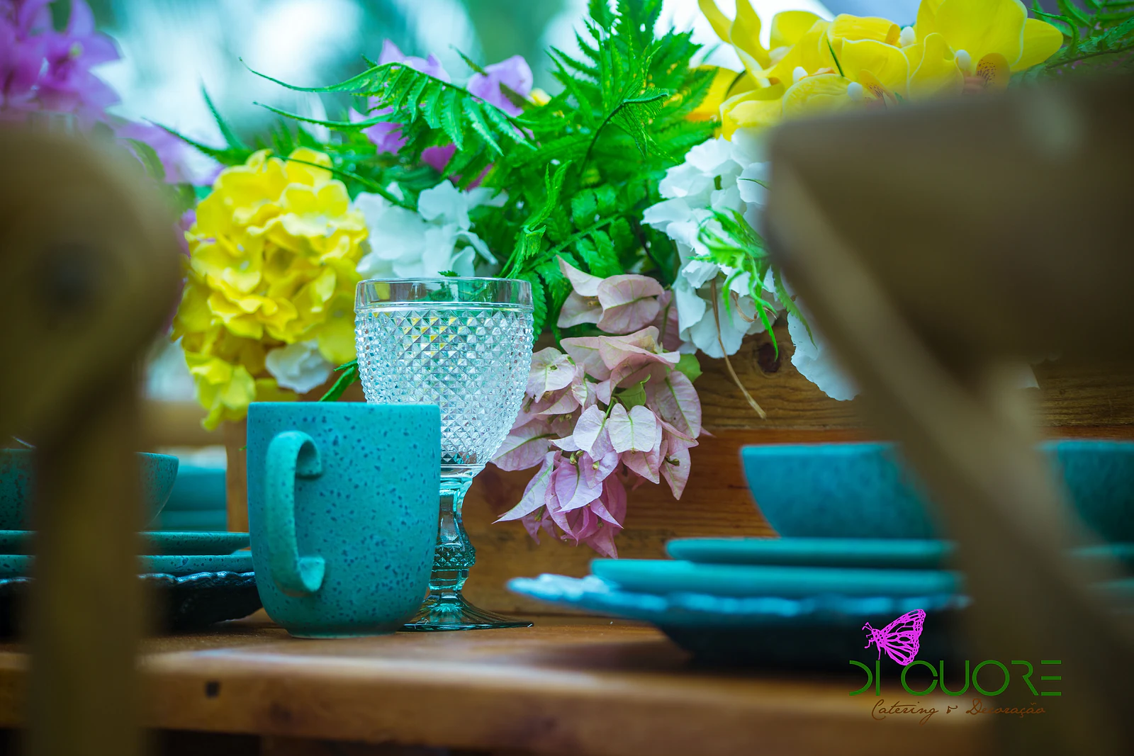 Caneca turquesa e copo de vidro lapidado ao lado de flores amarelas, brancas e rosa, vistos entre os encostos das cadeiras