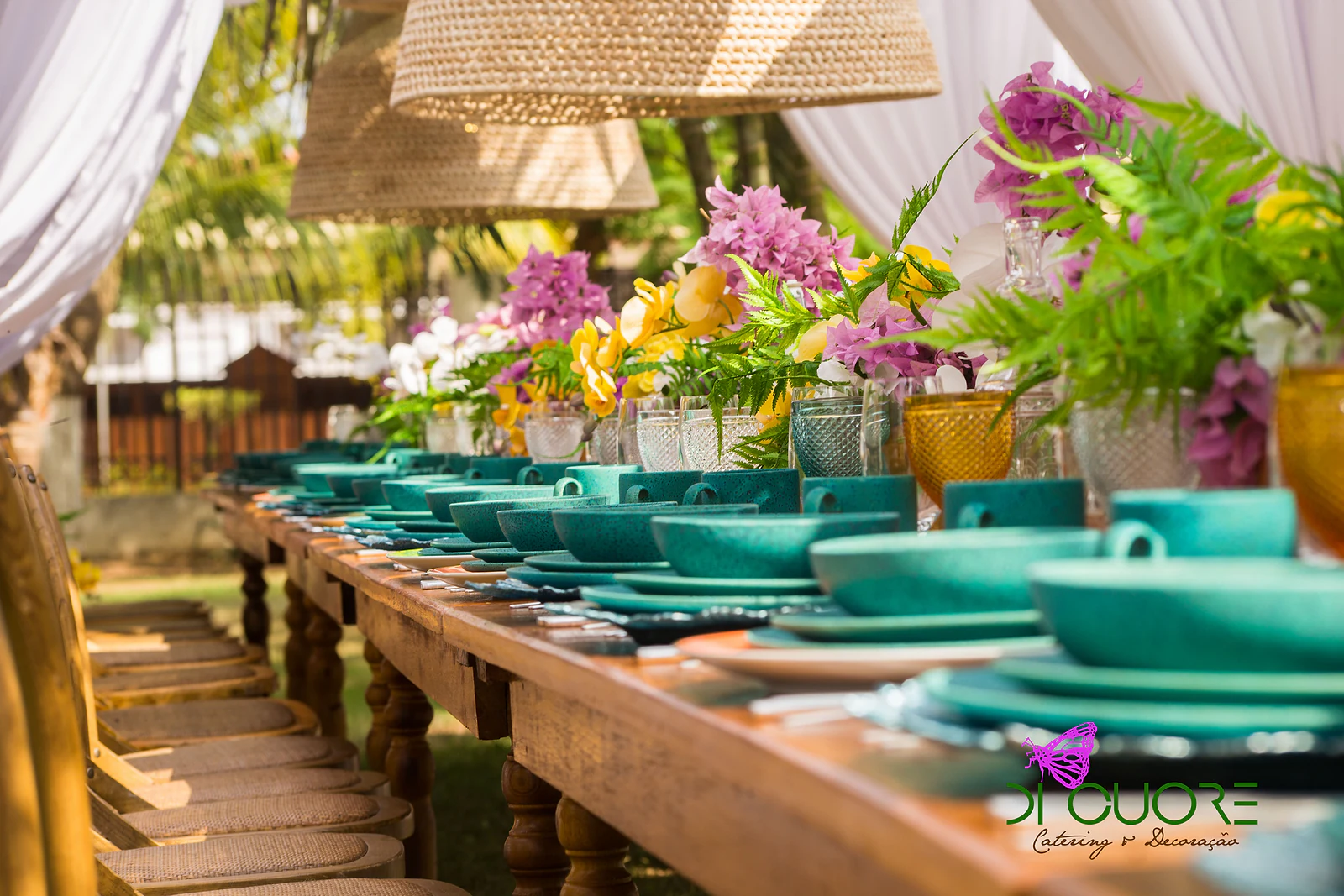 Mesa corrida de madeira com louça turquesa, copos lapidados e arranjos de orquídeas amarelas, buganvília lilás e fetos, sob candeeiros de palha