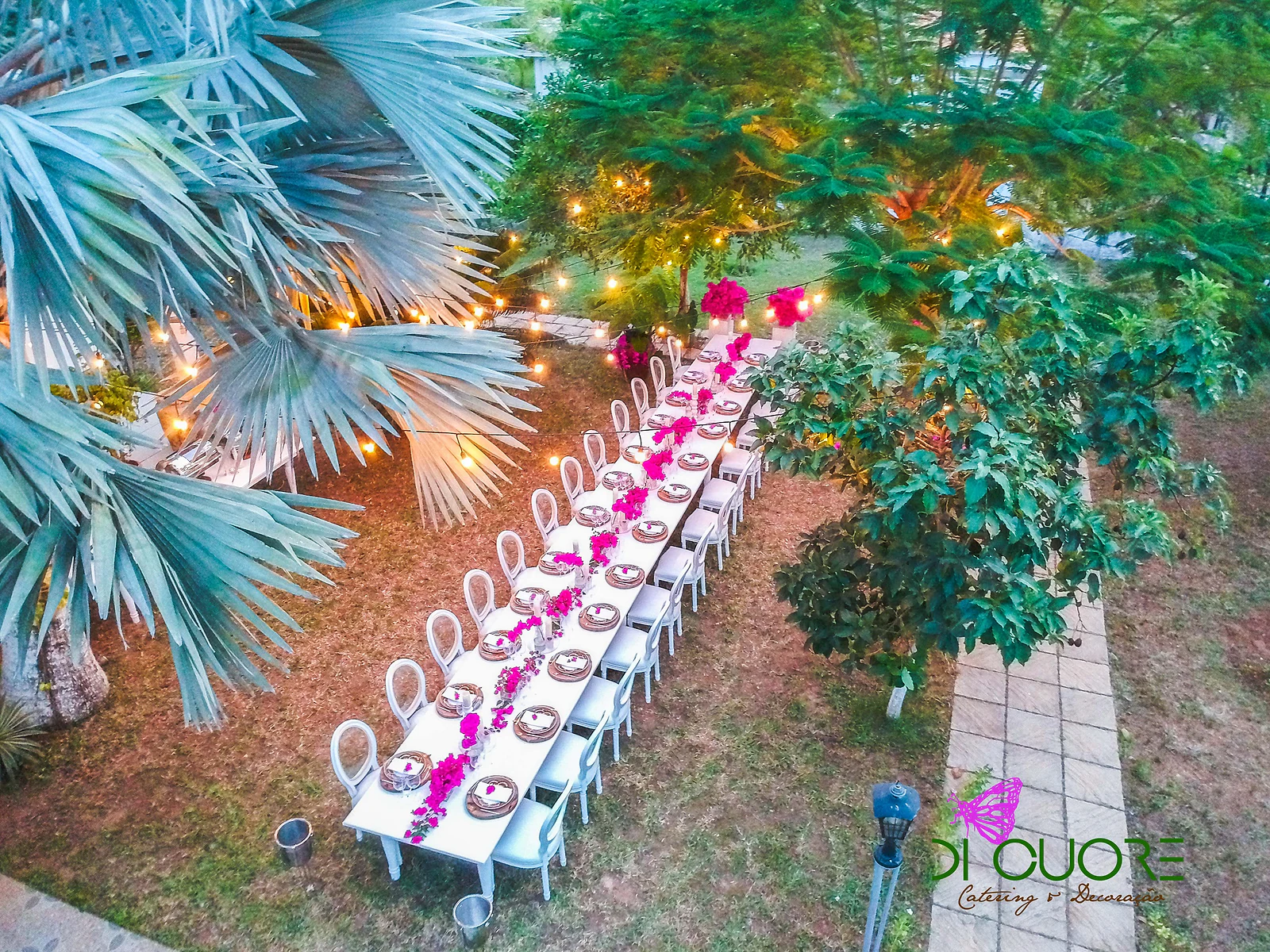 Aerial view of the long table among palms and trees, with white chairs and fuchsia flowers along its centre