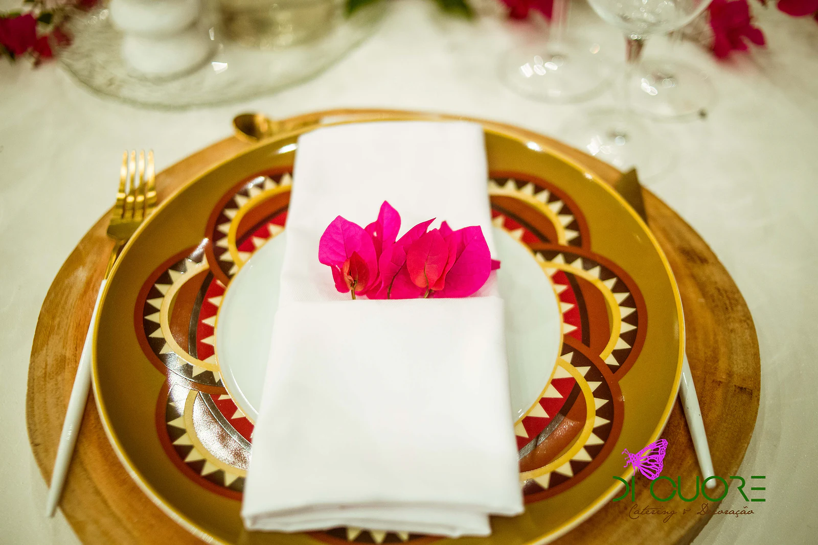 Plate with a geometric gold and earth-toned pattern on a wooden slice, with a white napkin and a bougainvillea bloom