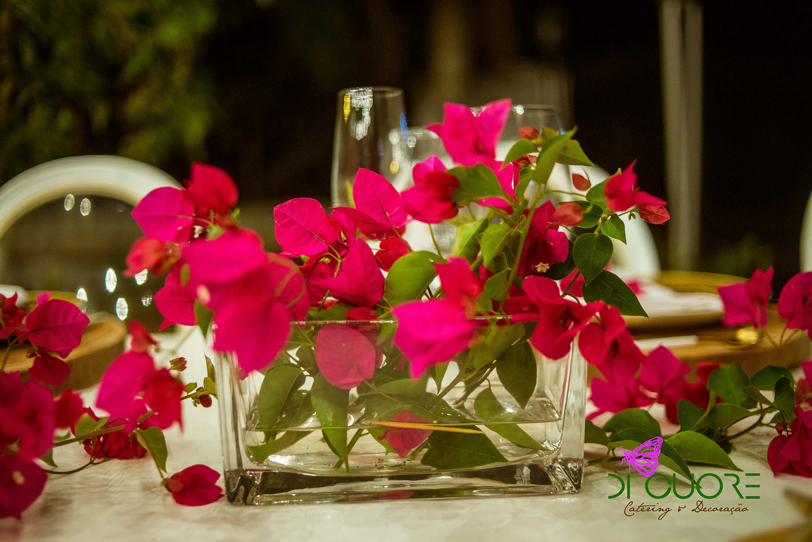 Rectangular glass vase of fuchsia bougainvillea sprigs on the table, with glasses behind