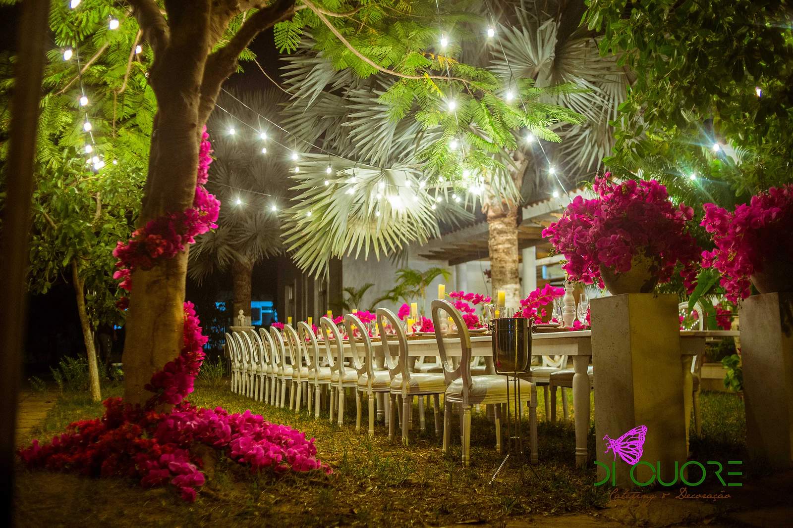 Long white table beneath lit trees, between pedestals with bougainvillea vases, a flower-wrapped trunk and petals strewn across the ground