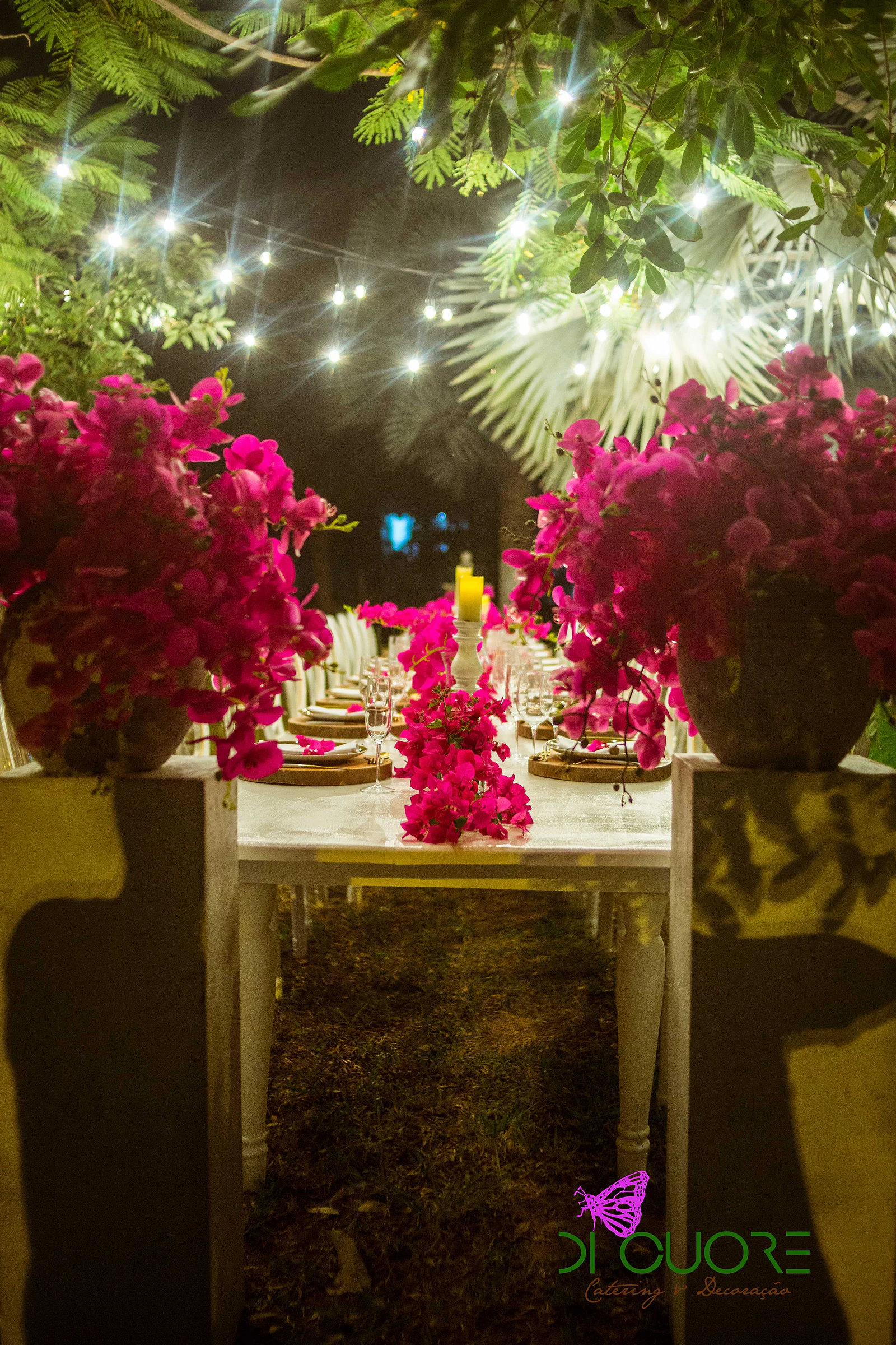 White table between two pedestals with large vases of fuchsia bougainvillea, beneath strings of lights and foliage