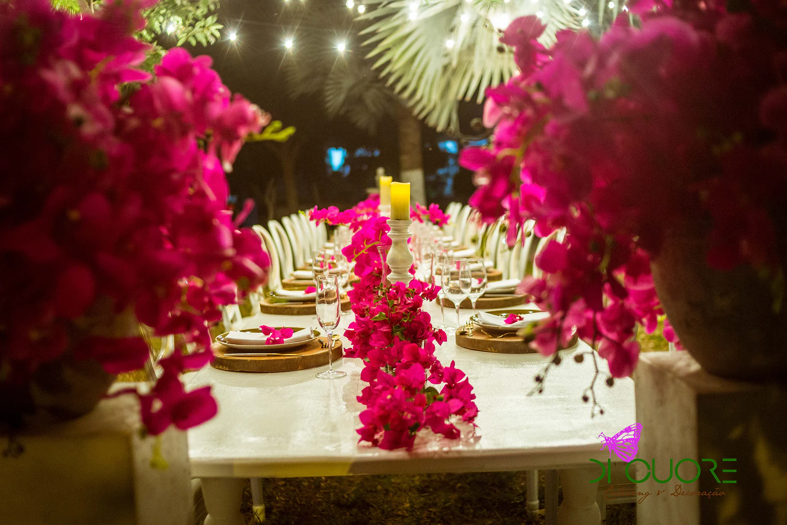 Mesa corrida de toalha branca com cadeiras brancas, velas e um caminho de buganvílias cor-de-rosa, sob luzes suspensas, num jantar ao ar livre à noite; em primeiro plano, ramos de buganvílias