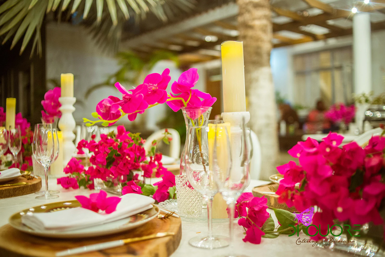 Side view of the table, with a row of white chairs, plates on wooden slices, yellow candles and sprays of fuchsia flowers