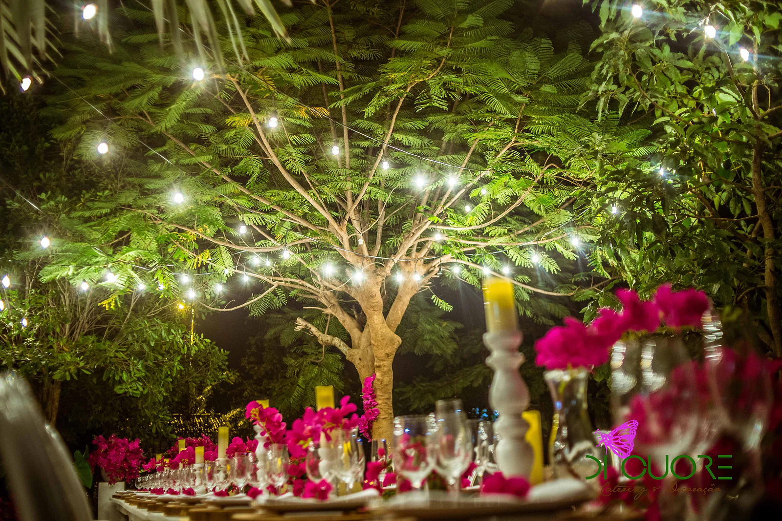Tree canopy lit by strings of lights above the laid table, with candles and fuchsia flowers in the foreground