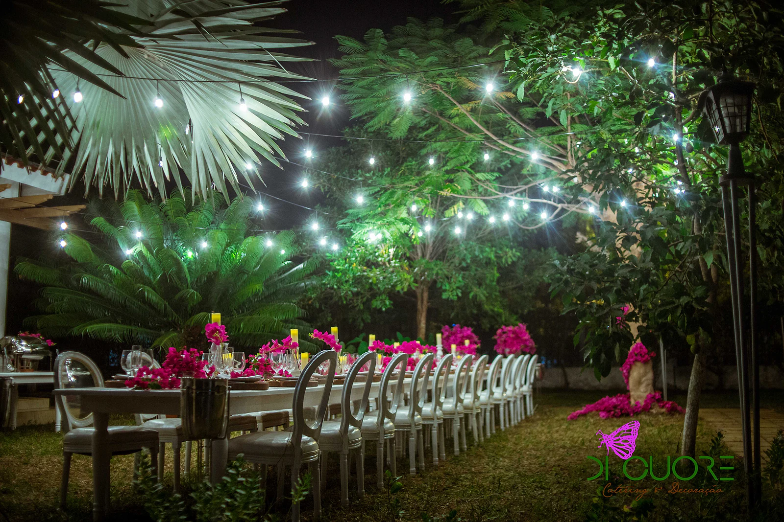 Long table at the far end of the lit garden, with fuchsia bougainvillea, petals on the lawn and trees hung with lights