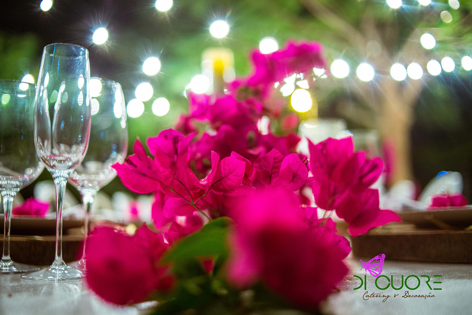 Close-up of fuchsia bougainvillea in the foreground, with crystal glasses and soft-focus lights behind