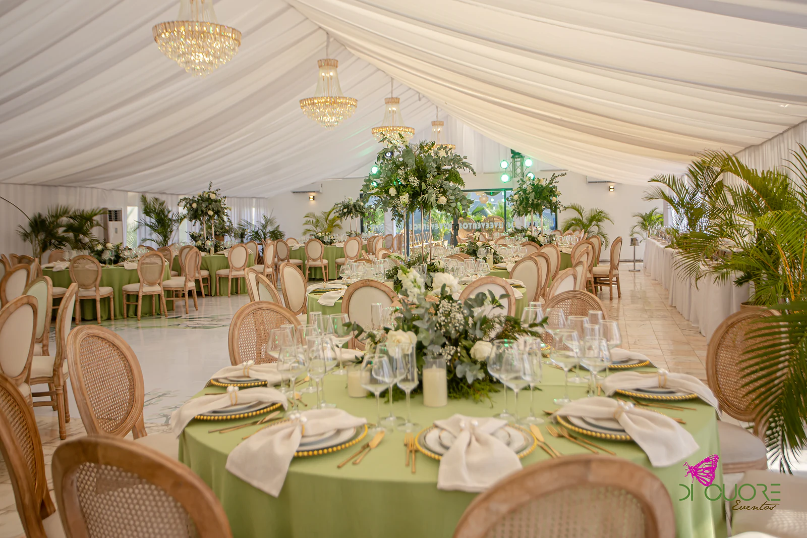 Mesa redonda de toalha verde-pistácio com centro de eucalipto e rosas brancas, sob candelabros de cristal e panejamento branco
