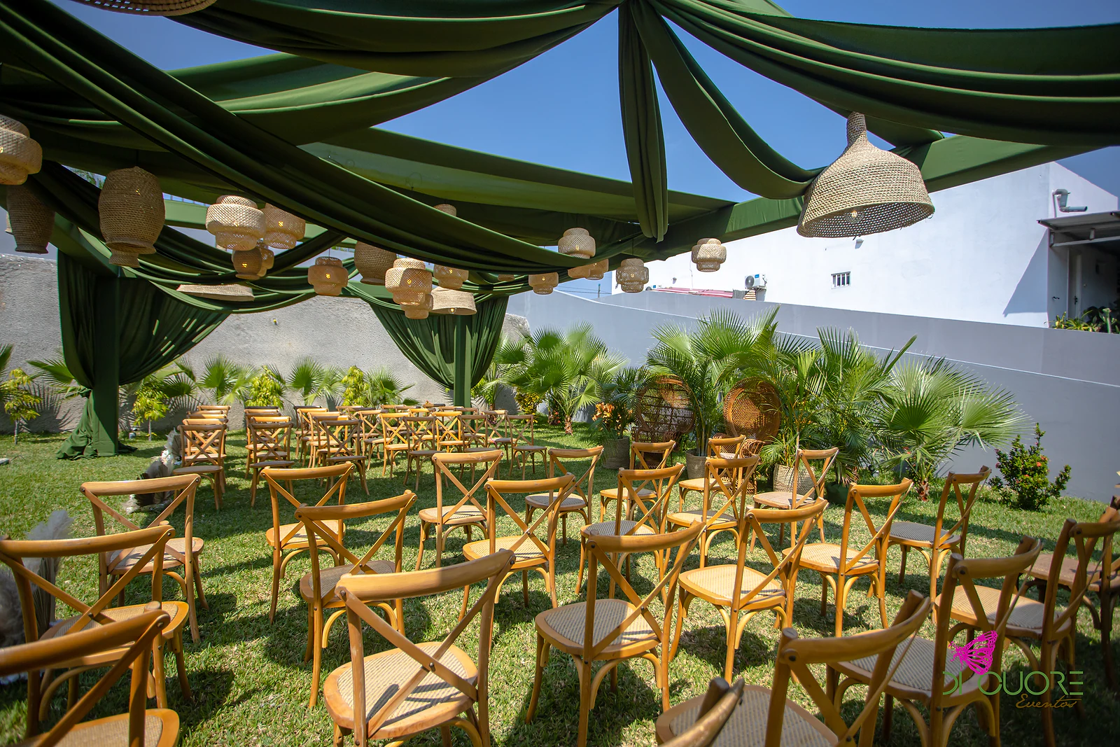 Fileiras de cadeiras de madeira sobre a relva, sob uma pérgola vestida de cortinados verdes e lanternas de fibra natural, com palmeiras e um muro branco ao fundo