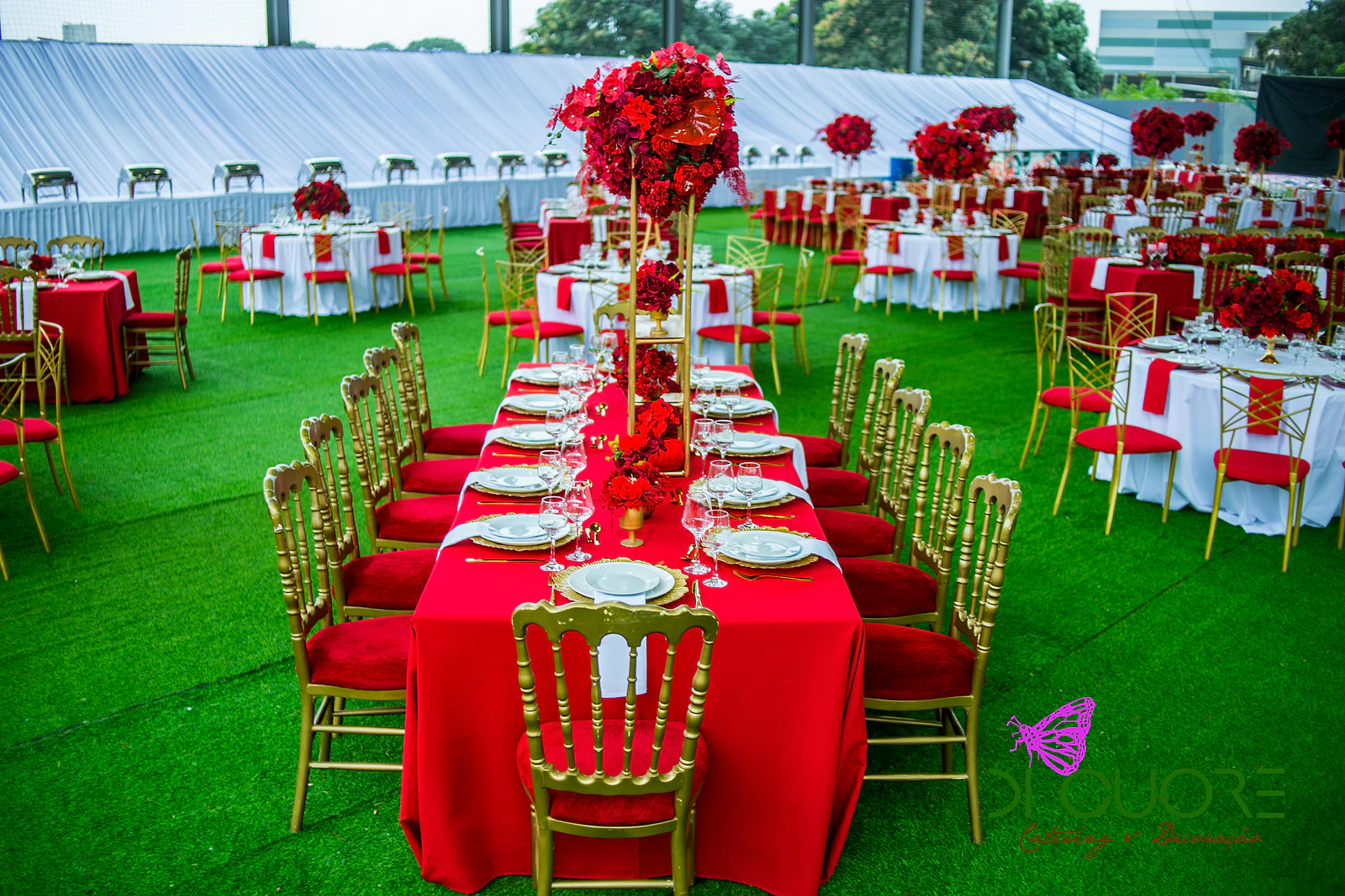 Mesa comprida de toalha vermelha com cadeiras douradas de almofada vermelha e um centro de mesa alto de rosas, sobre a relva