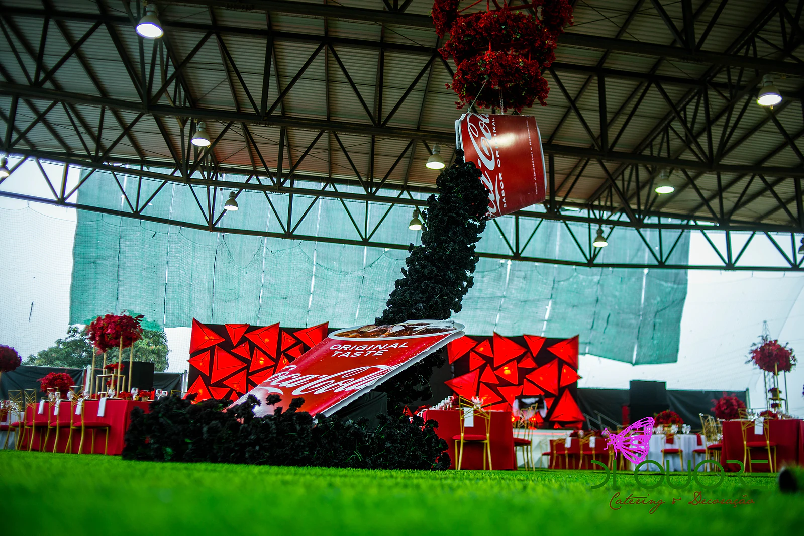 Lata suspensa de um aro de flores vermelhas a despejar uma cascata de rosas negras sobre a relva, com o palco vermelho e mesas ao fundo