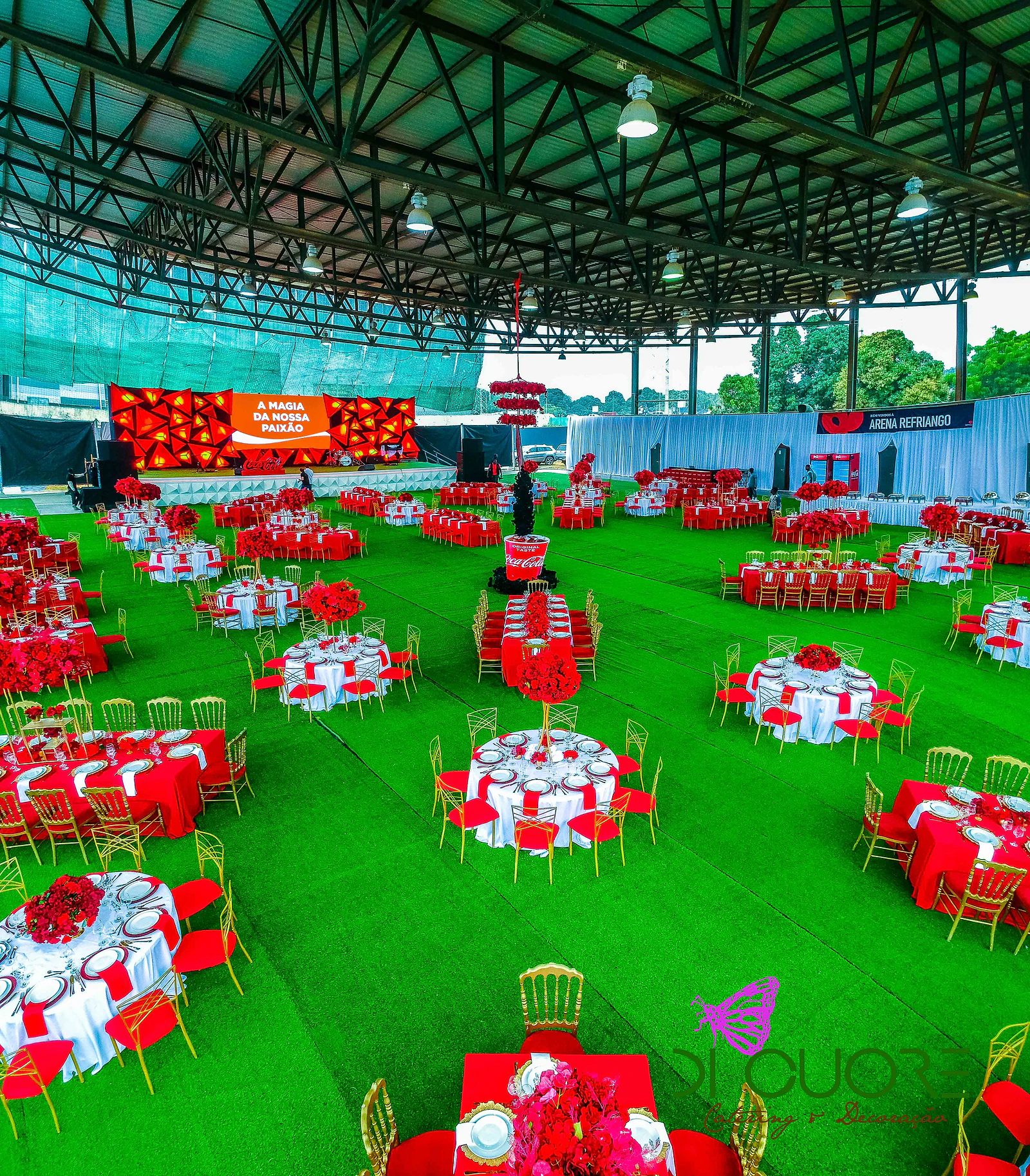 Vista geral da cobertura com mesas redondas e compridas de toalhas vermelhas e brancas, cadeiras douradas, relva verde e o palco ao fundo