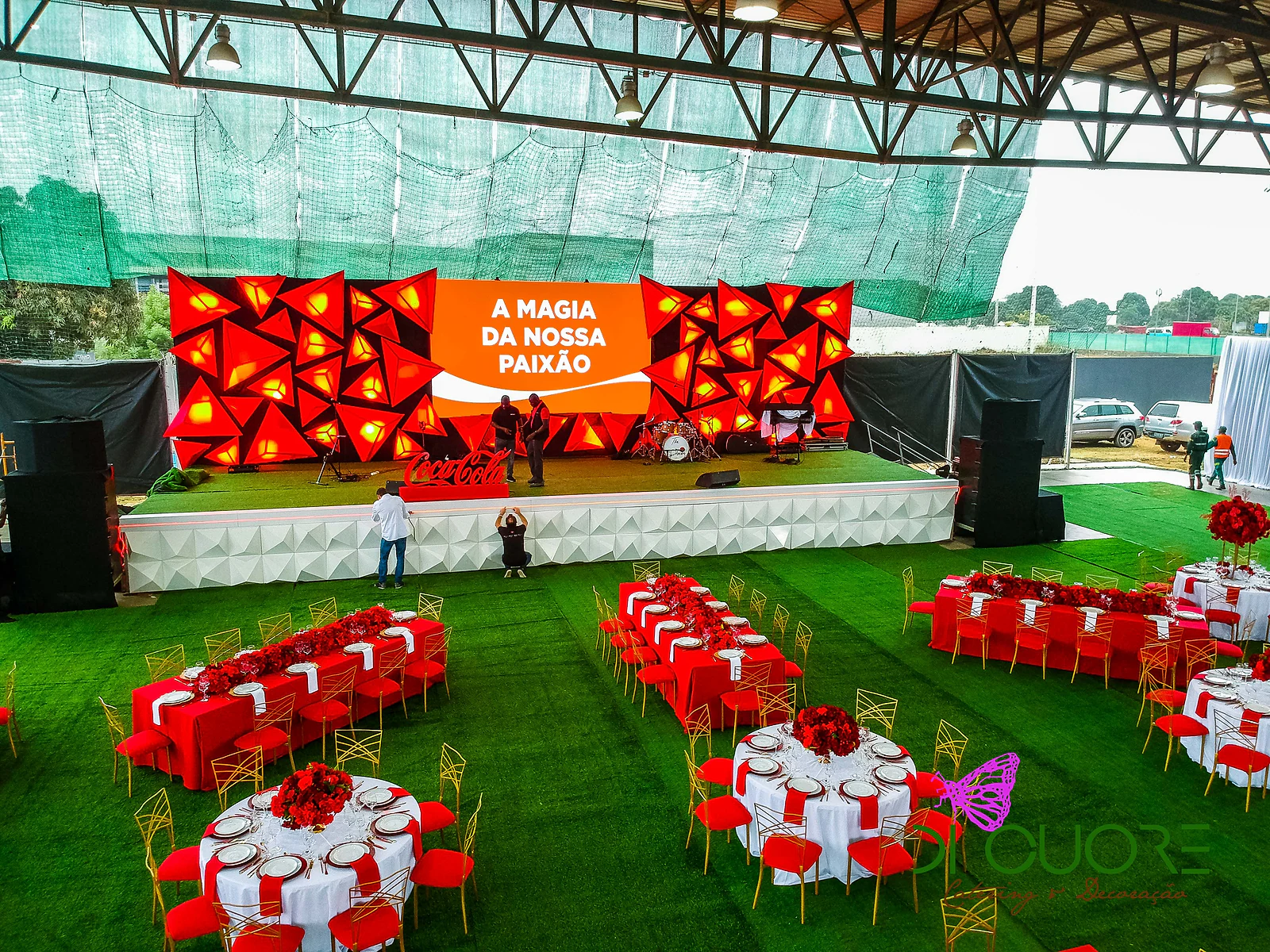Palco com fundo de triângulos vermelhos e o lema “A Magia da Nossa Paixão”, sobre uma base branca facetada, com mesas redondas e compridas à frente