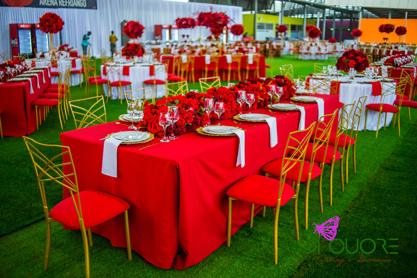 Mesa de toalha vermelha com um friso de rosas vermelhas ao centro, subpratos dourados, guardanapos brancos e cadeiras douradas