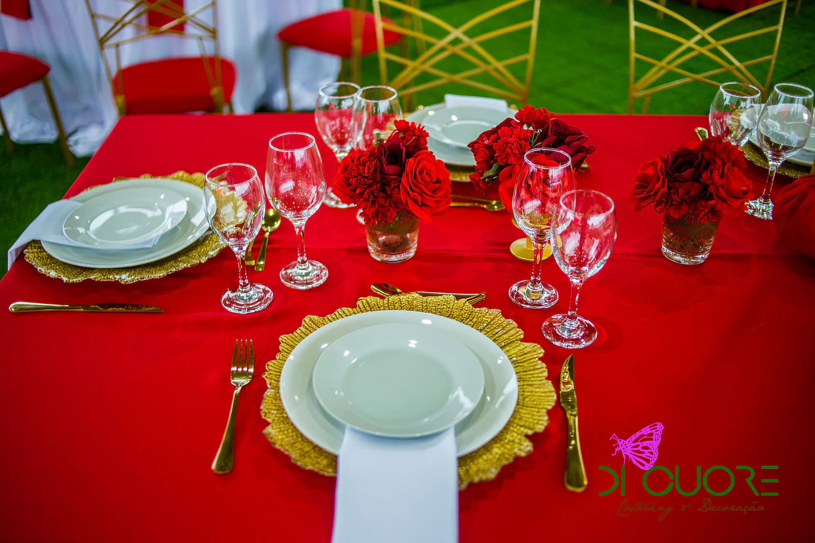 Lugar à mesa com subprato dourado, prato branco, talheres dourados e copos, entre pequenos ramos de rosas vermelhas, sobre toalha vermelha