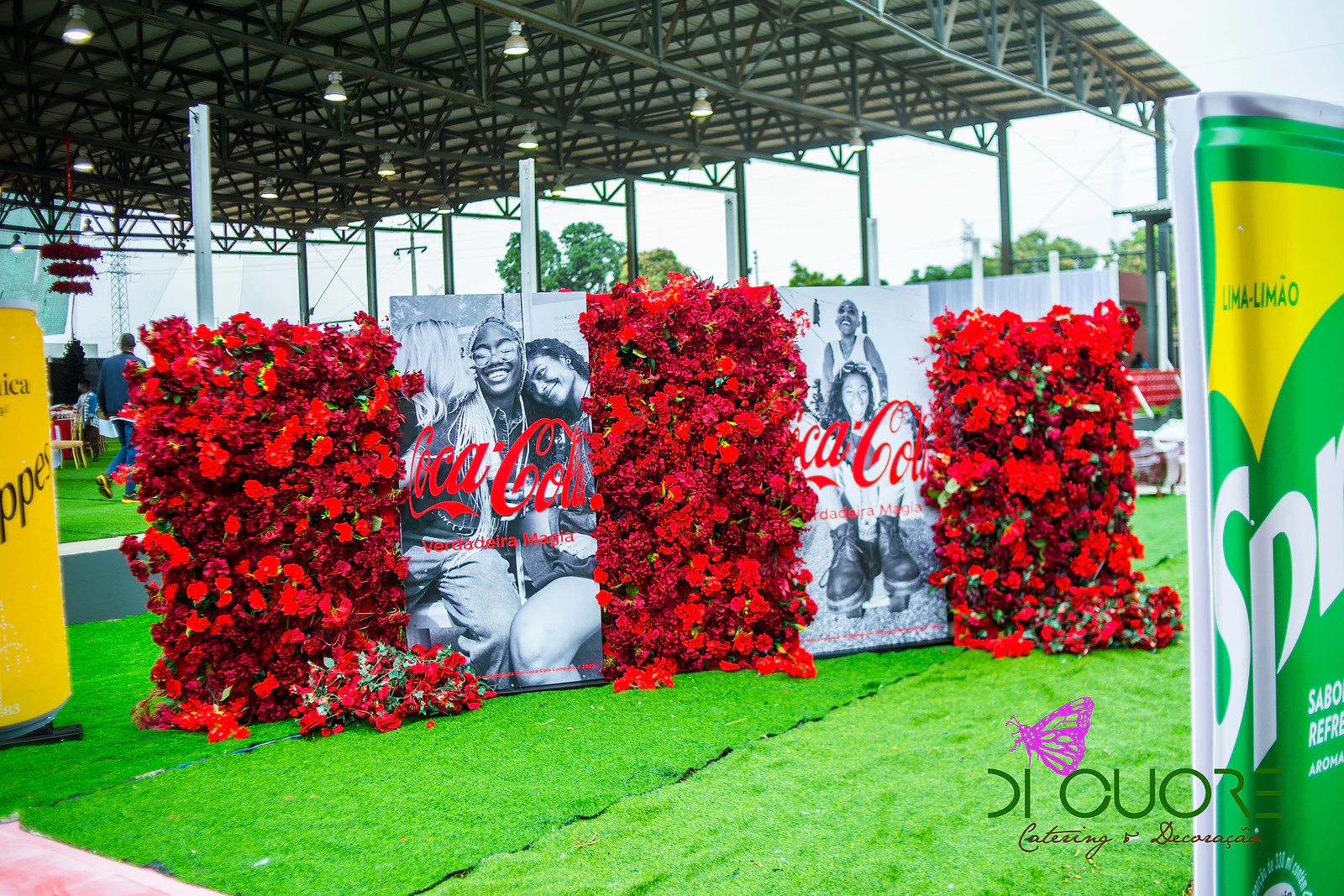Parede de rosas vermelhas com painéis de fotografias a preto e branco, entre garrafas e latas gigantes, sobre relva