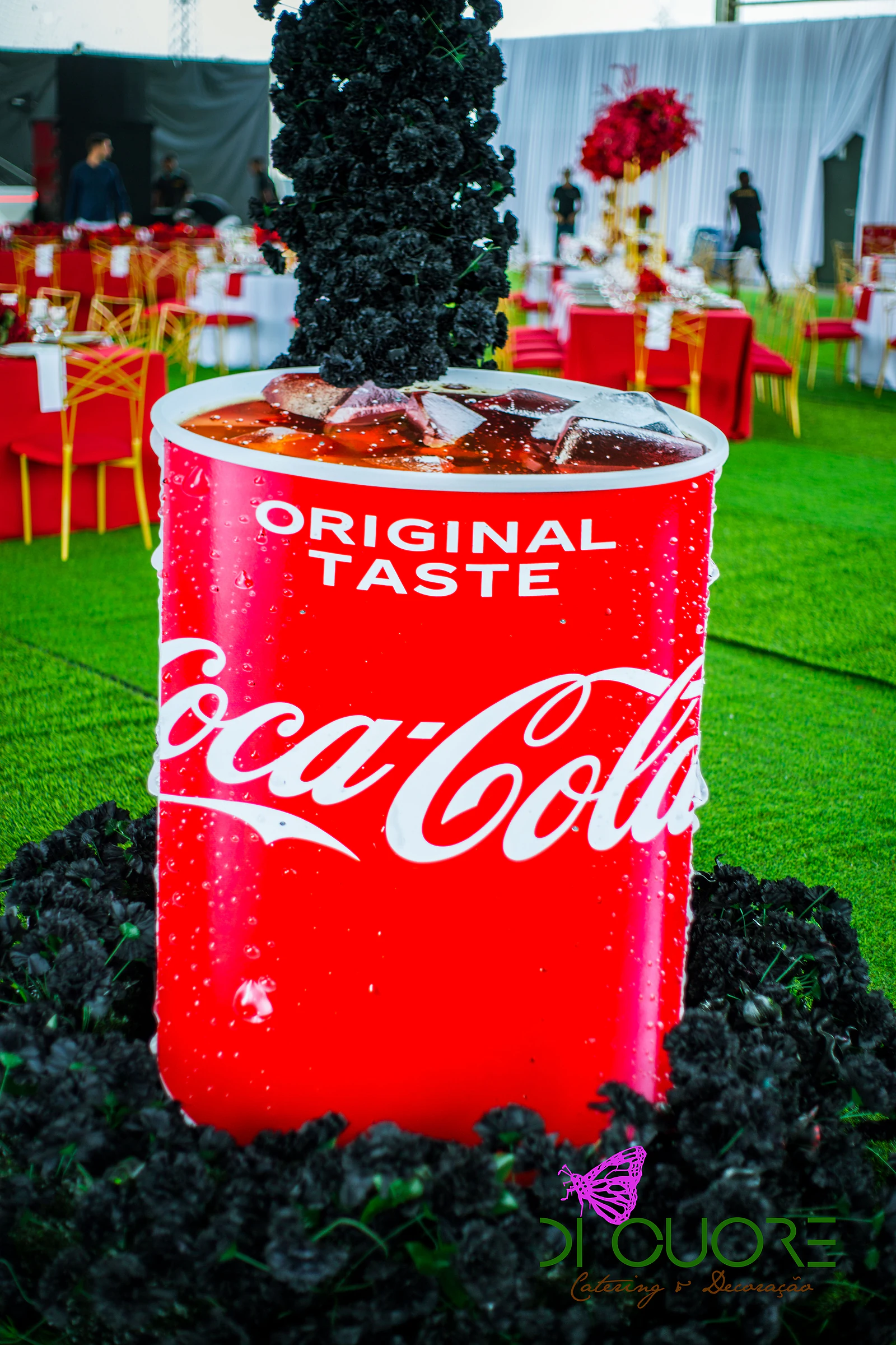 Copo gigante com a marca em vermelho e gelo à boca, com uma coluna de rosas negras a cair e um anel de rosas negras à volta, sobre a relva
