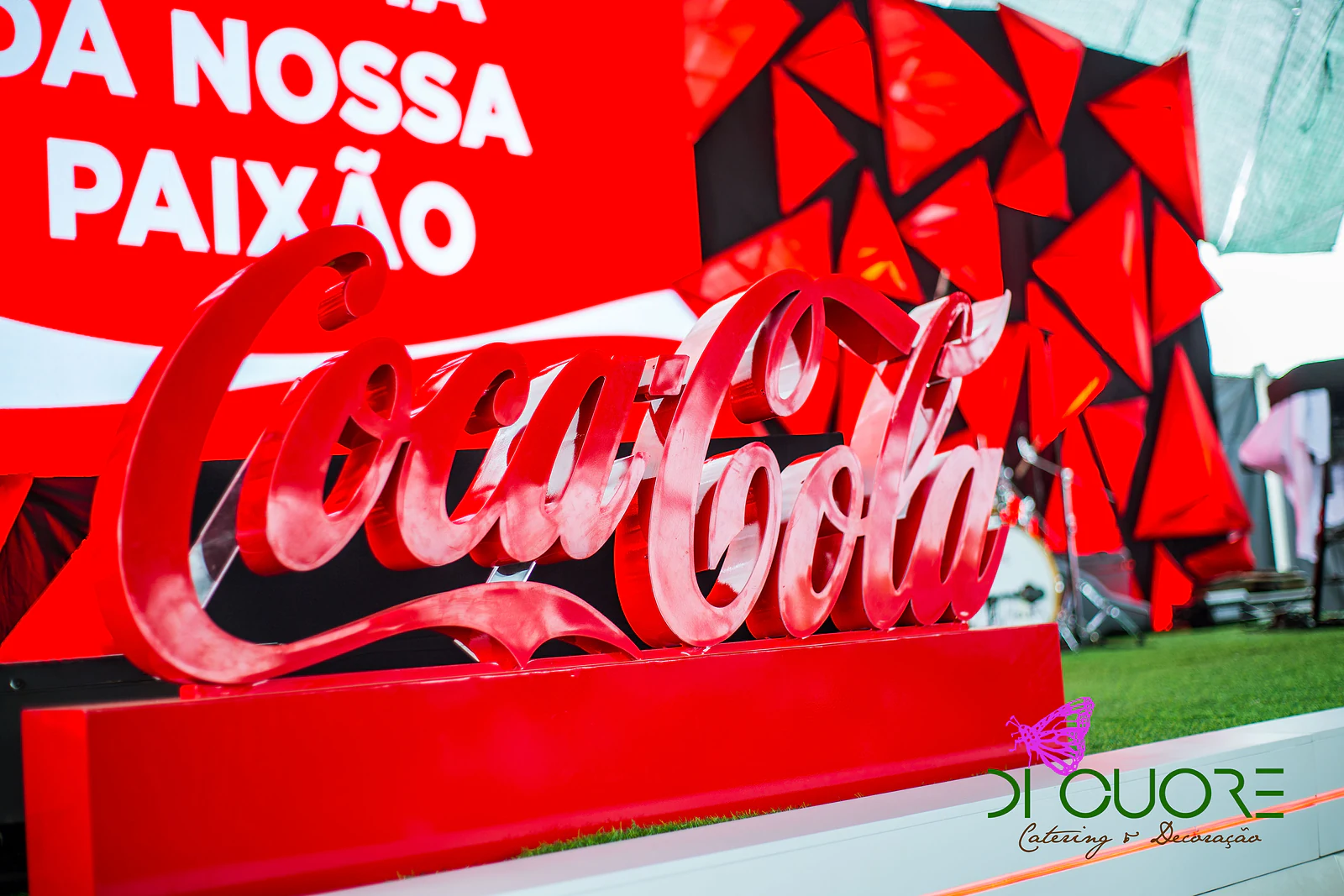 Letras vermelhas do logótipo da Coca-Cola sobre um pedestal vermelho, com o painel e os triângulos vermelhos do palco ao fundo