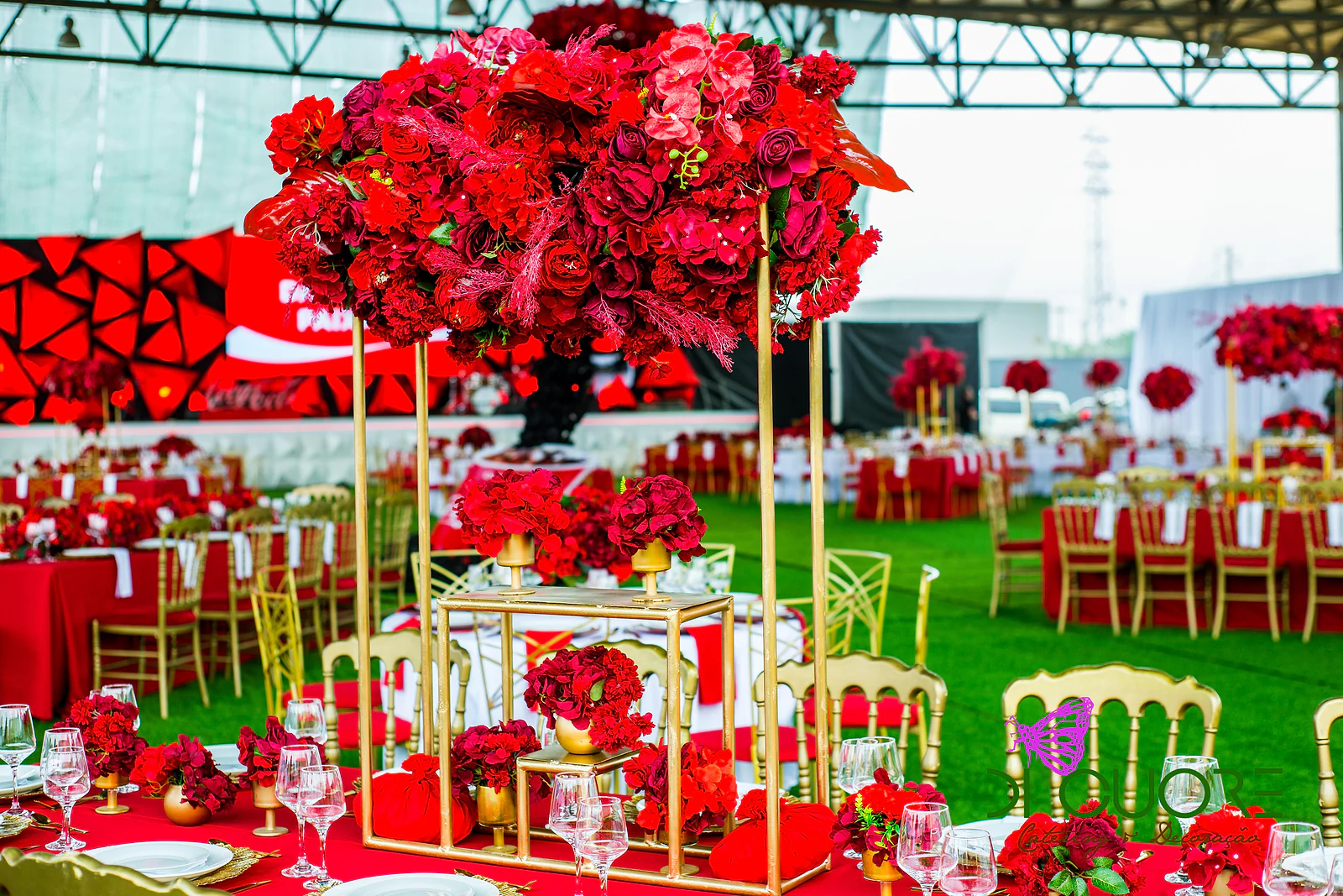 Tall arrangement of red flowers on a gold frame, with tables laid in red and gold chairs on turf, in a hall with red panels behind
