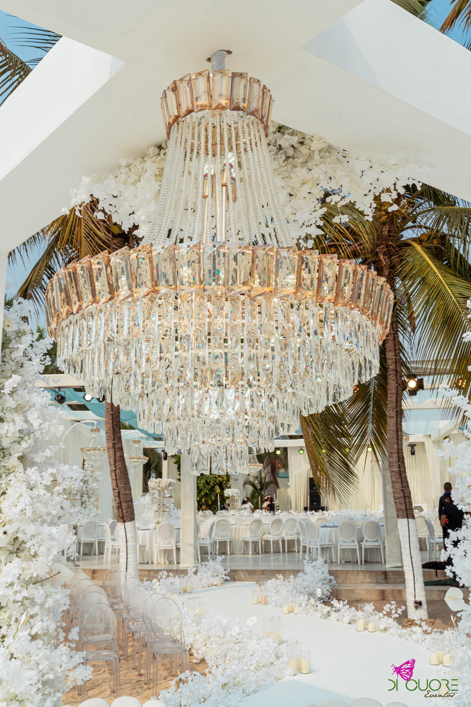 Lustre de cristal dourado sob o arco branco, com palmeiras e o corredor de cadeiras transparentes ao fundo