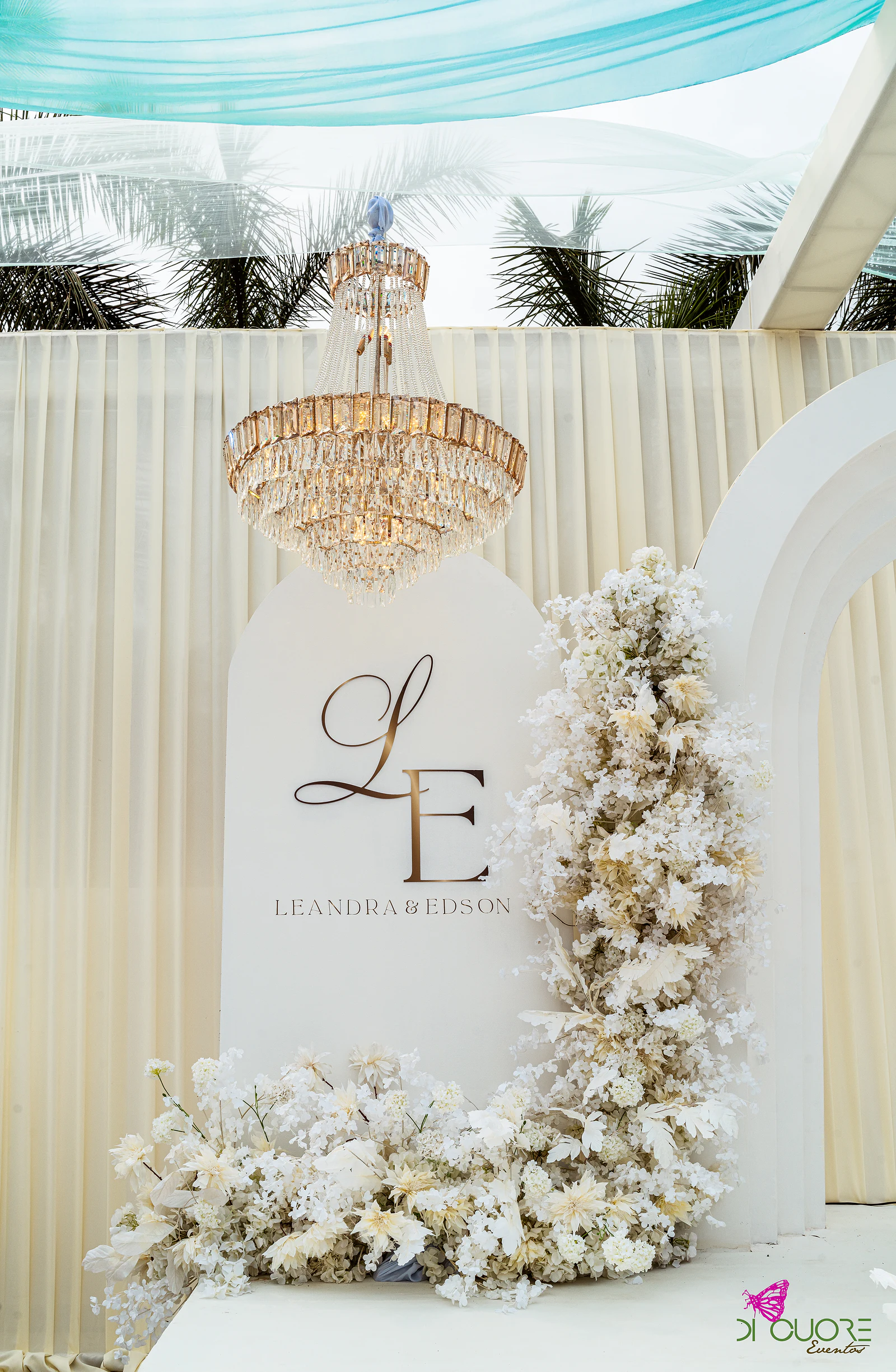 Painel com o monograma L&E sob um lustre de cristal, ao lado de uma coluna de flores brancas e cor de creme
