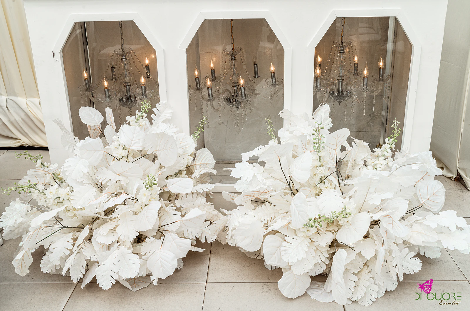 Móvel branco com três nichos de espelho e candeeiros de cristal lá dentro, ladeado por grandes arranjos de folhas e flores brancas