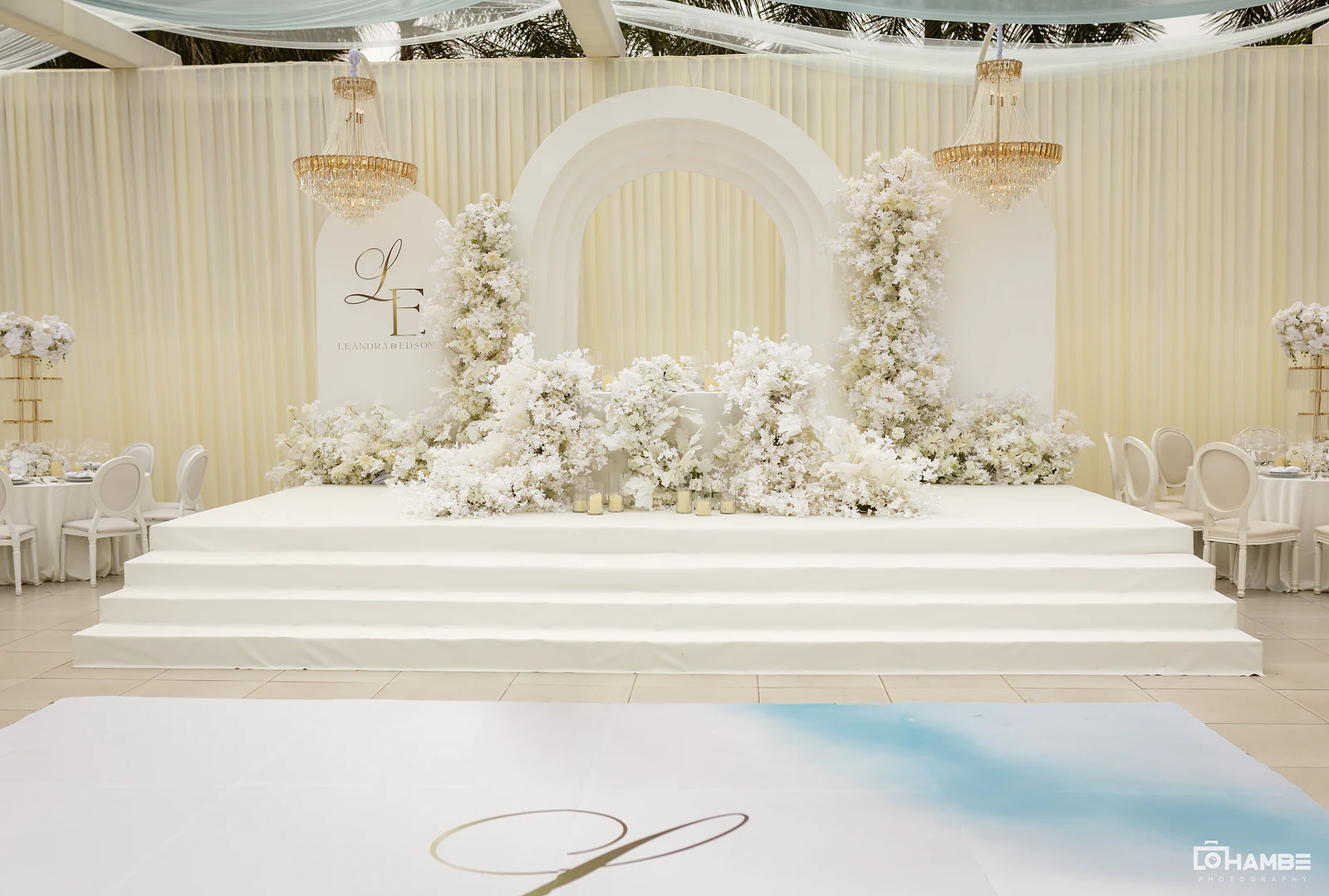 Palco de degraus brancos com o arco, o painel L&E e colunas de flores brancas entre dois lustres dourados, e a pista com o monograma em primeiro plano
