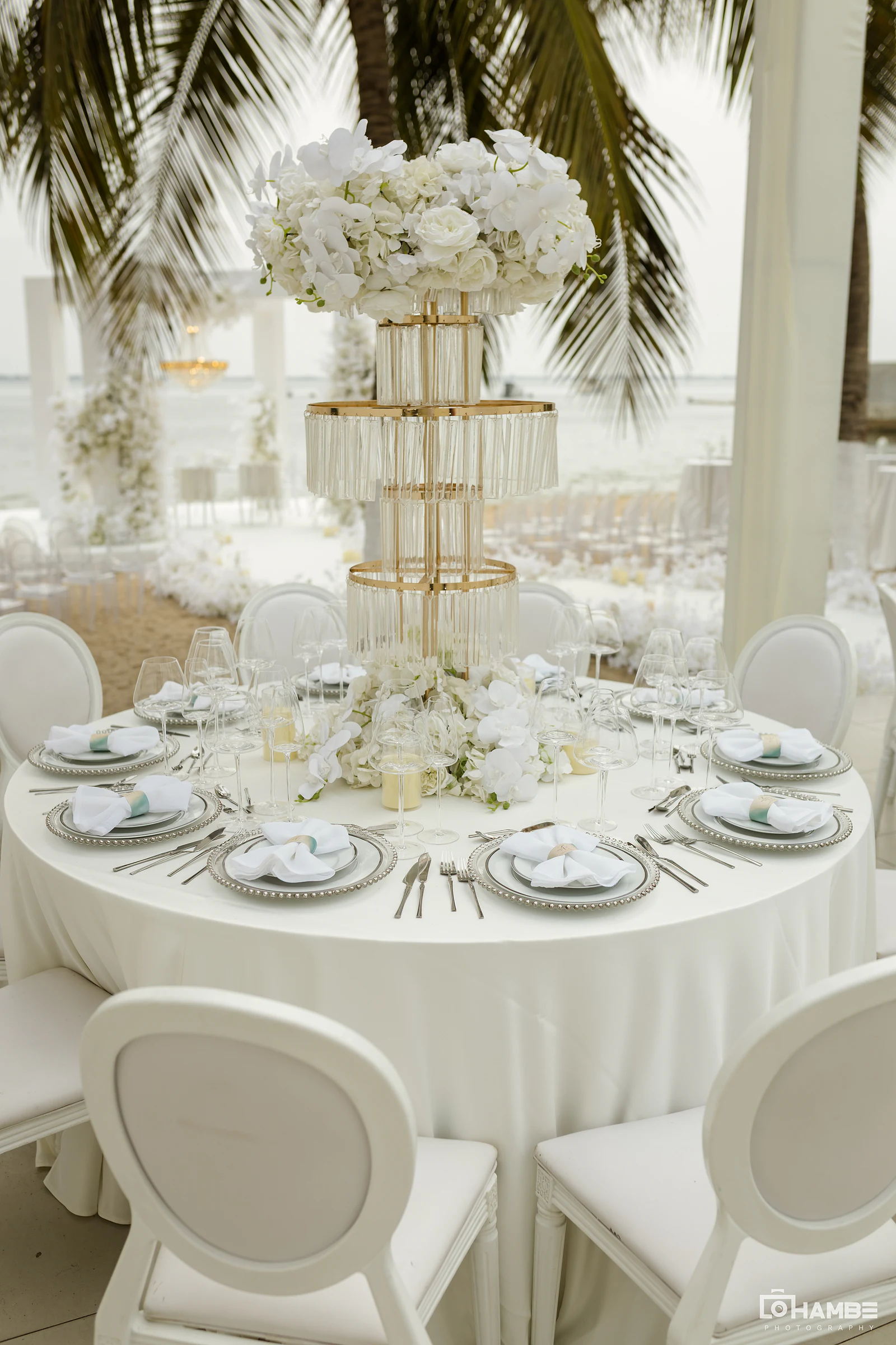 Mesa redonda de linho branco com centro de mesa dourado e de cristal, rematado por flores brancas, pratos de rebordo de contas e cadeiras brancas, com o mar atrás