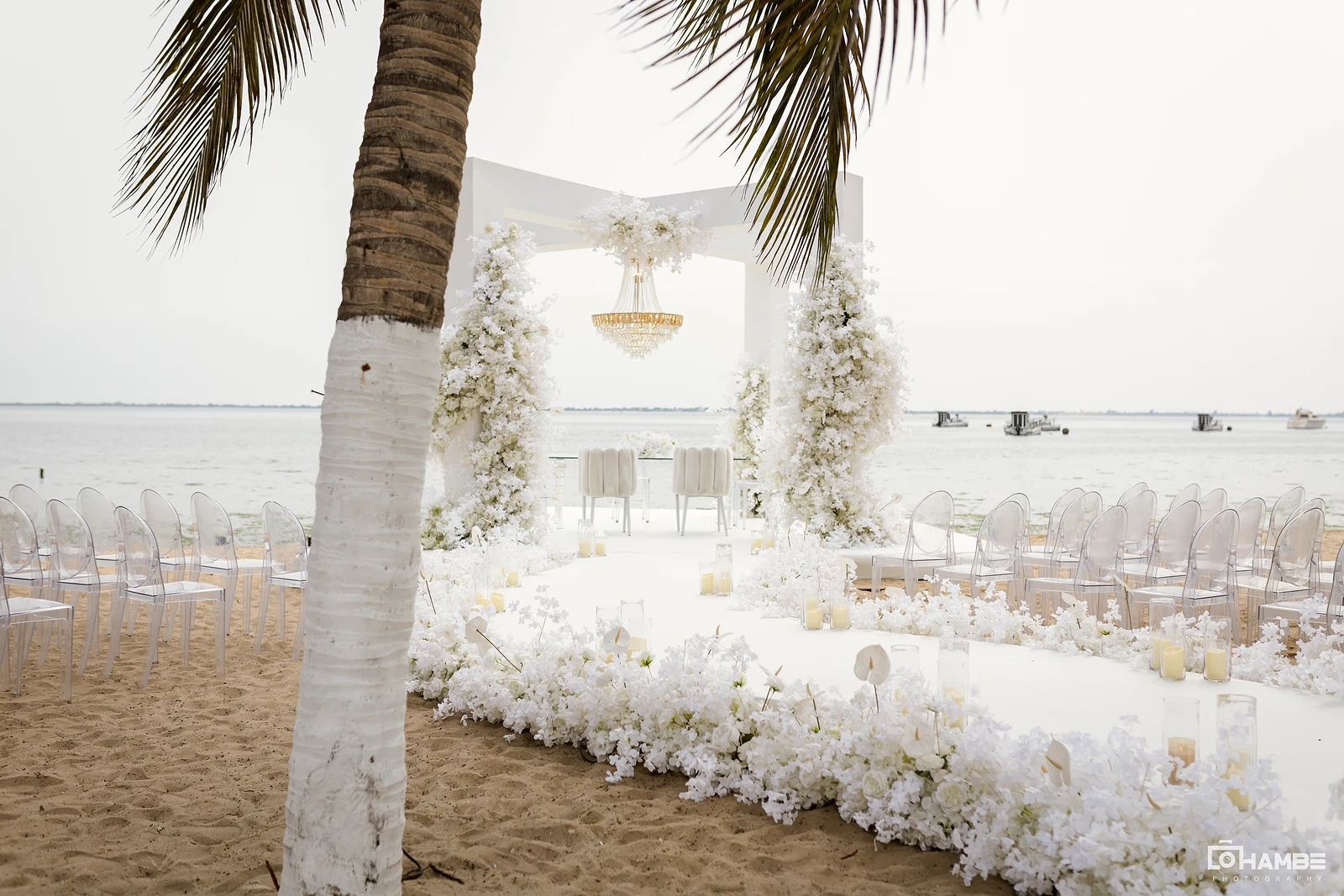 Passadeira branca em curva entre flores e velas até ao arco com lustre, com cadeiras transparentes na areia e o mar ao fundo