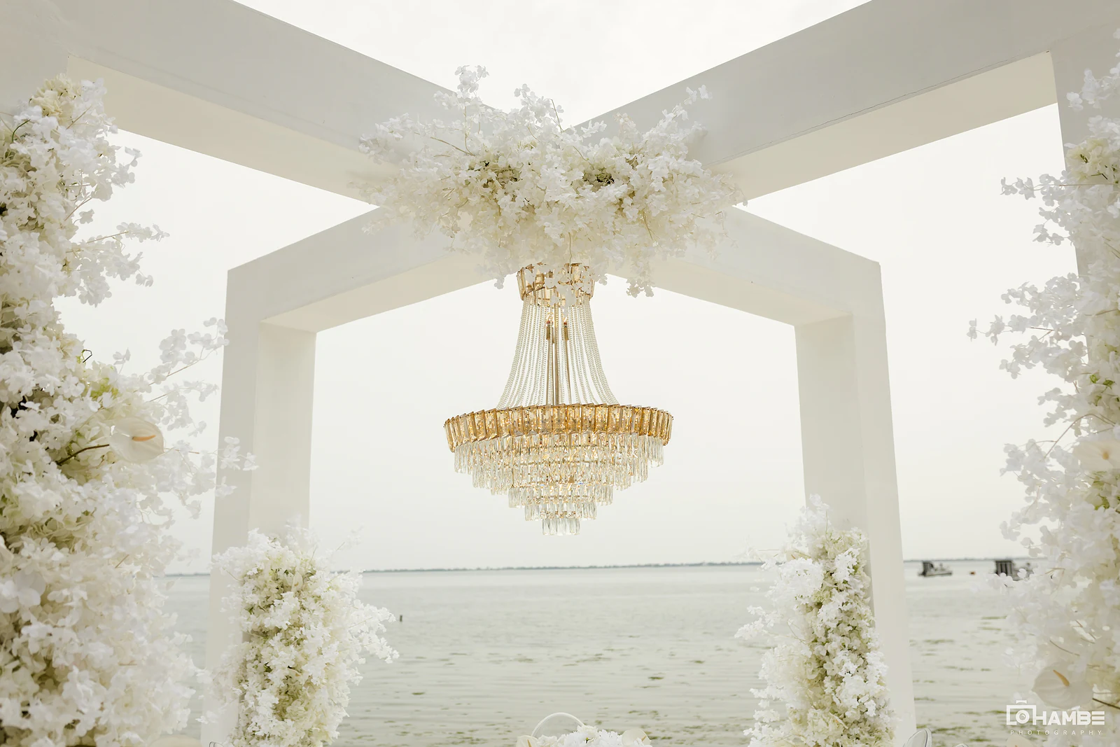Arco branco com lustre de cristal e flores brancas, com o mar ao fundo